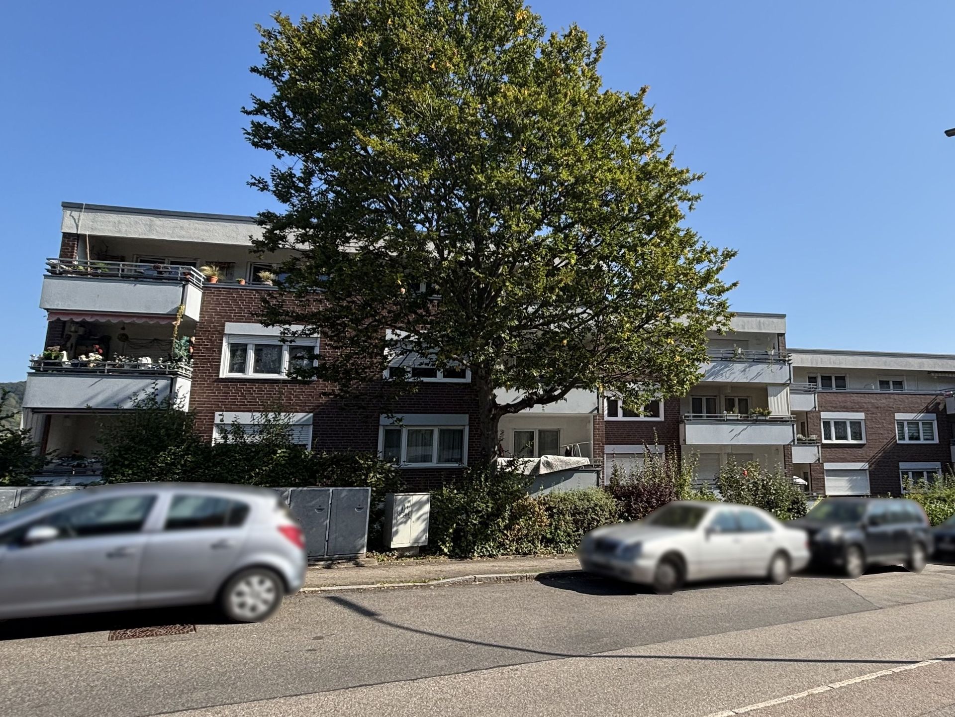 Das Bild zeigt ein Mehrfamilienhaus mit brauner Fassade und mehreren Balkonen. Vor dem Haus stehen einige Bäume und parkende Autos am Straßenrand.