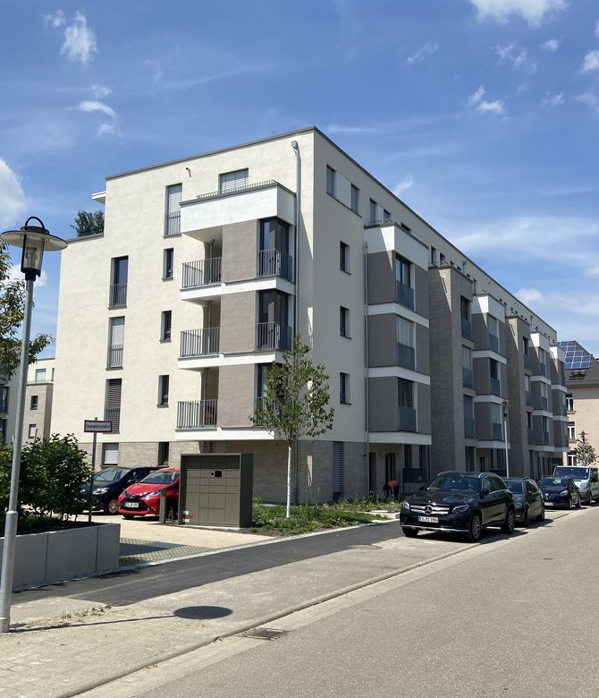 Das Bild zeigt ein Mehrfamilienhaus mit weißer Fassade und grauen Elementen. Vor dem Haus sind Autos geparkt. Link zum Bereich Neubau Eigentum.