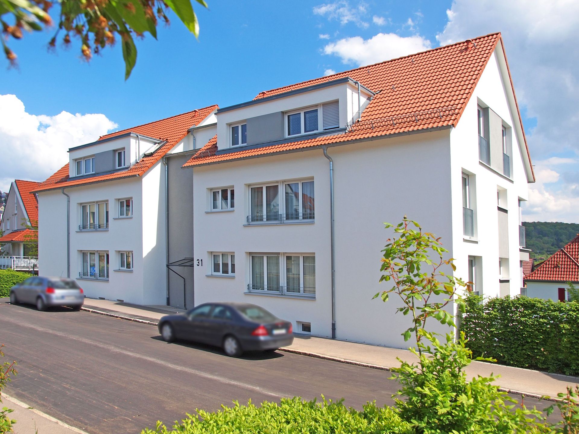 Das Bild zeigt zwei moderne Mehrfamilienhäuser mit weißer Fassade, roten Ziegeldächern und großen Fenstern. Die Häuser stehen an einer ruhigen Straße mit geparkten Autos und sind von grünen Büschen umgeben.