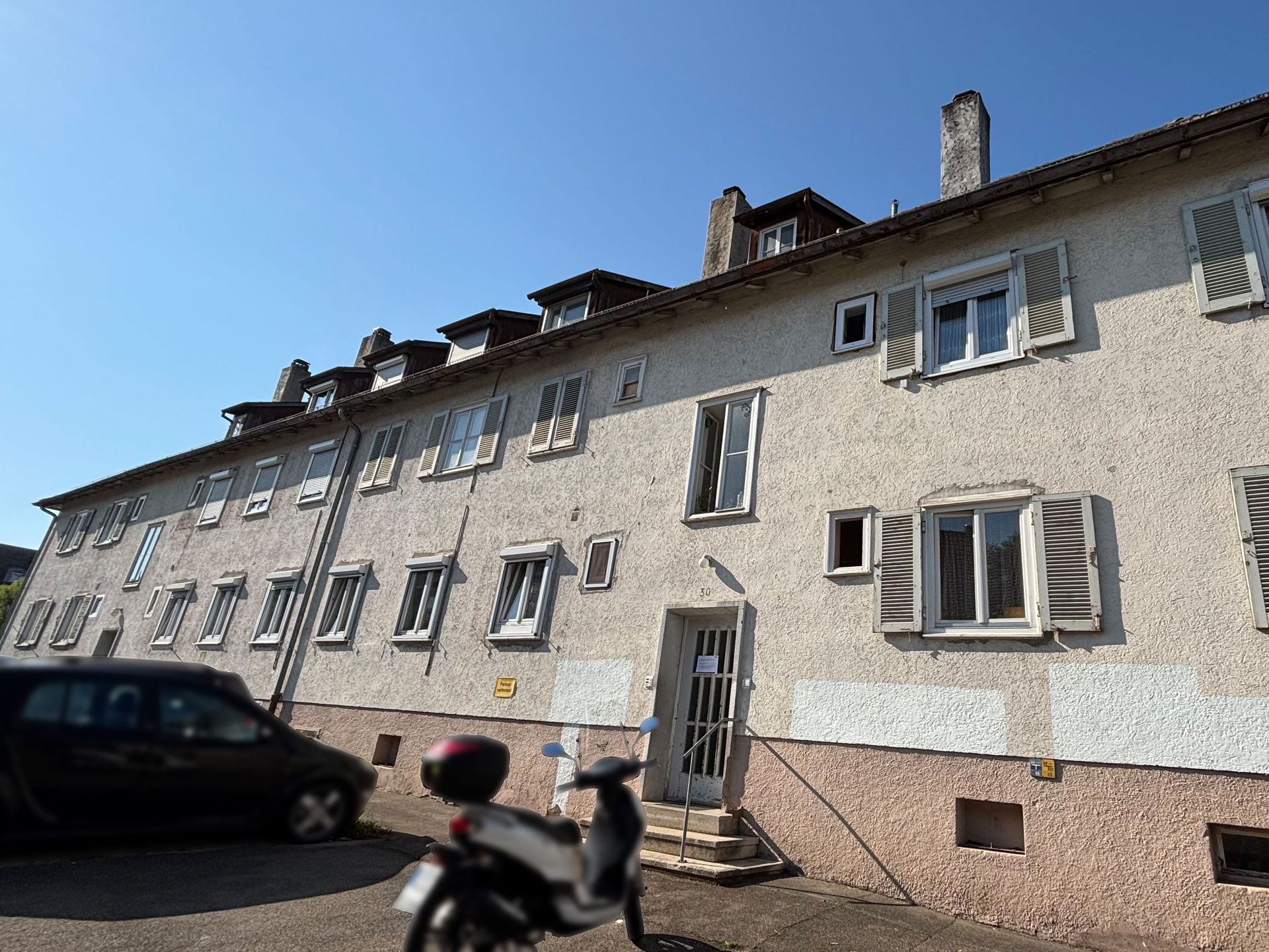 Das Bild zeigt ein langgezogenes Mehrfamilienhaus mit heller Fassade und Dachgauben.  Die Fenster sind mit weißen Fensterläden ausgestattet und vor dem Haus befinden sich ein geparktes Auto und ein Motorroller.