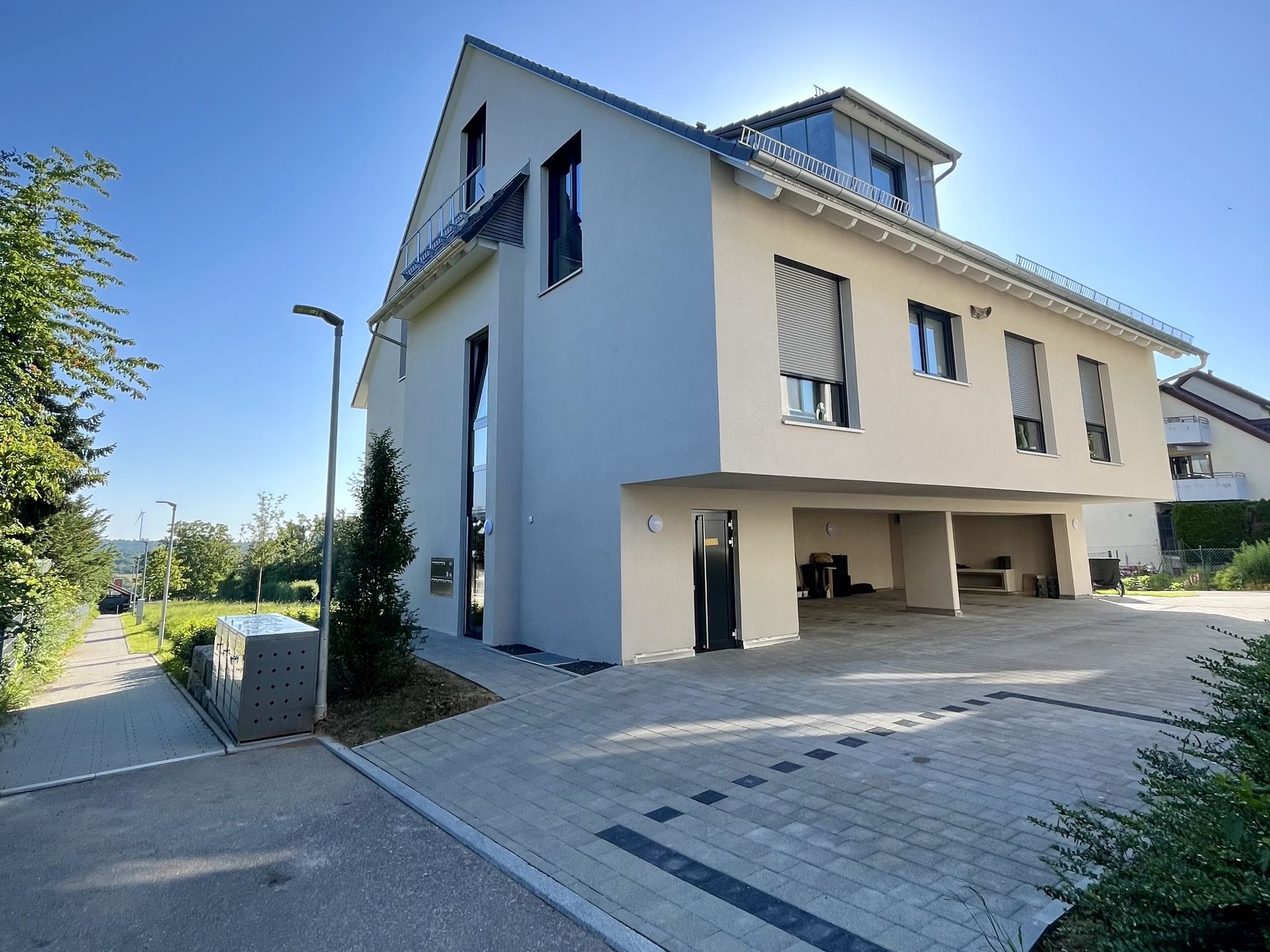 Das Bild zeigt ein Mehrfamilienhaus mit heller Fassade und einer gepflasterten Fläche vor dem Haus.