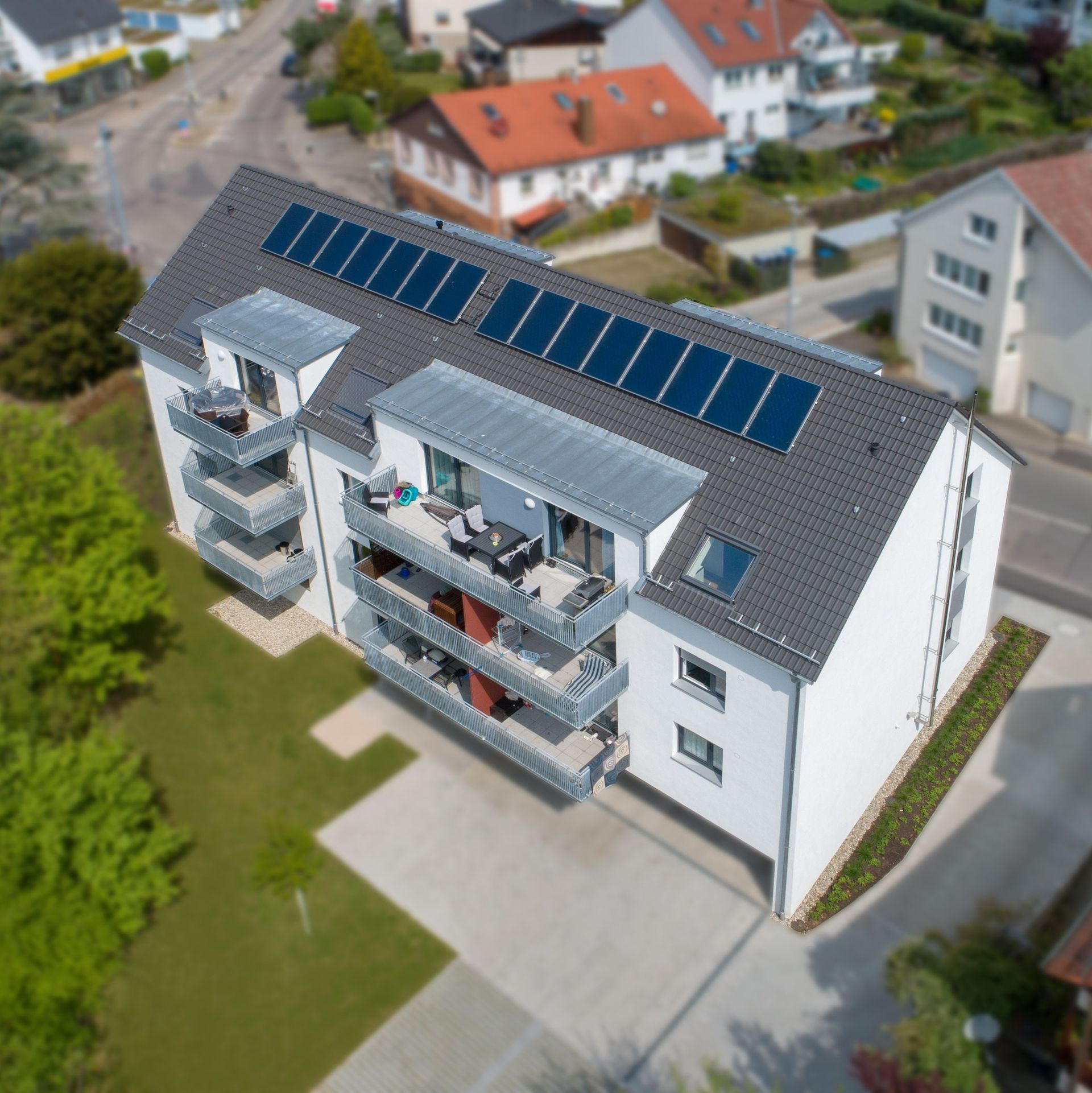 Die Luftaufnahme zeigt ein modernes Mehrfamilienhaus mit grauen Dachziegeln und Solaranlage. Das Gebäude hat mehrere Balkone und ist von Grünflächen umgeben.