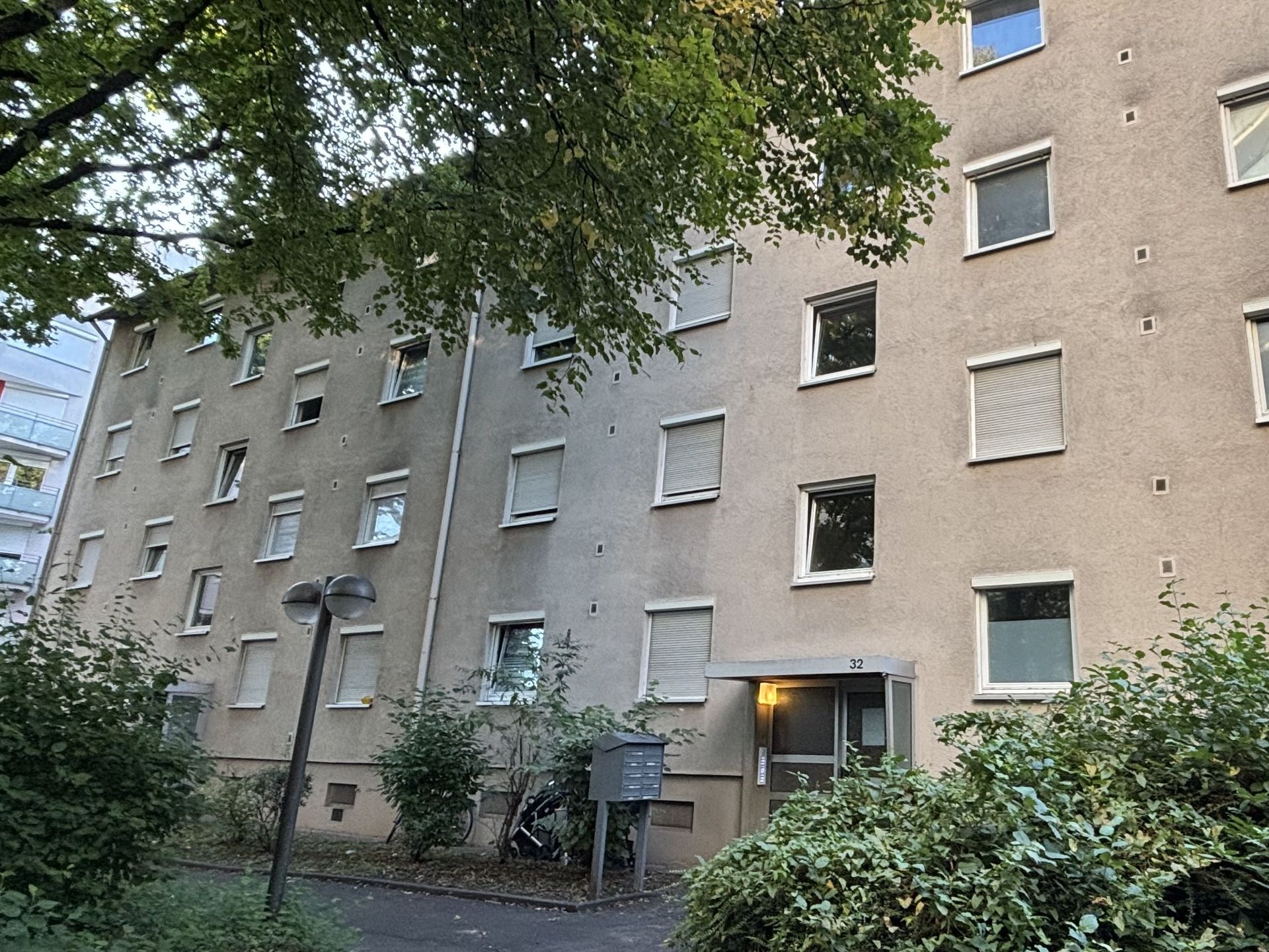 Das Bild zeigt ein mehrstöckiges Wohngebäude mit heller Fassade und vielen Fenstern, die mit Rollläden versehen sind.  Vor dem Haus stehen einige Bäume und Büsche, und ein gepflasterter Weg führt zum Eingang.