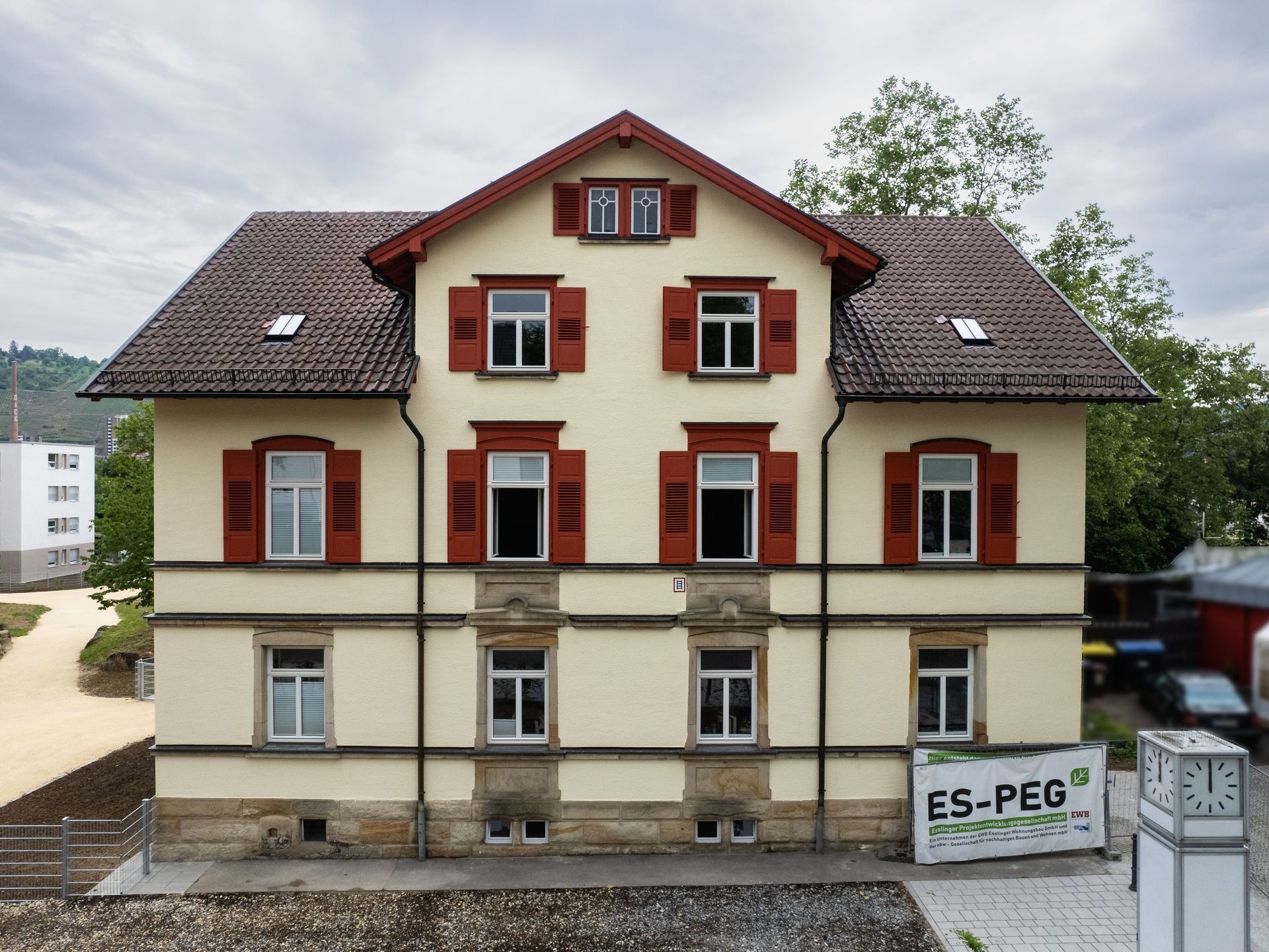 Das Bild zeigt ein Gebäude mit heller Fassade, braunem Dach und braunen Fensterläden. Vor dem Haus befindet sich eine unbefestigte Fläche.