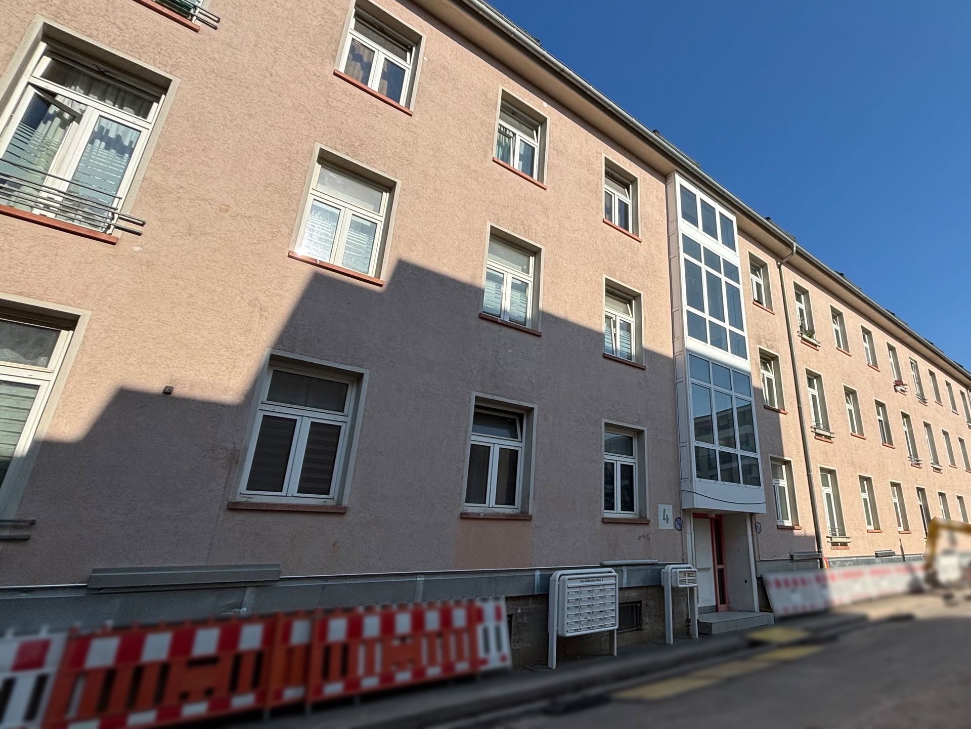 Das Bild zeigt ein Mehrfamilienhaus mit heller Fassade und vielen Fenstern, an dessen Seite ein moderner, verglaster Anbau mit Treppenhaus zu sehen ist. Vor dem Haus befindet sich eine Baustellenabsperrung und Briefkästen.