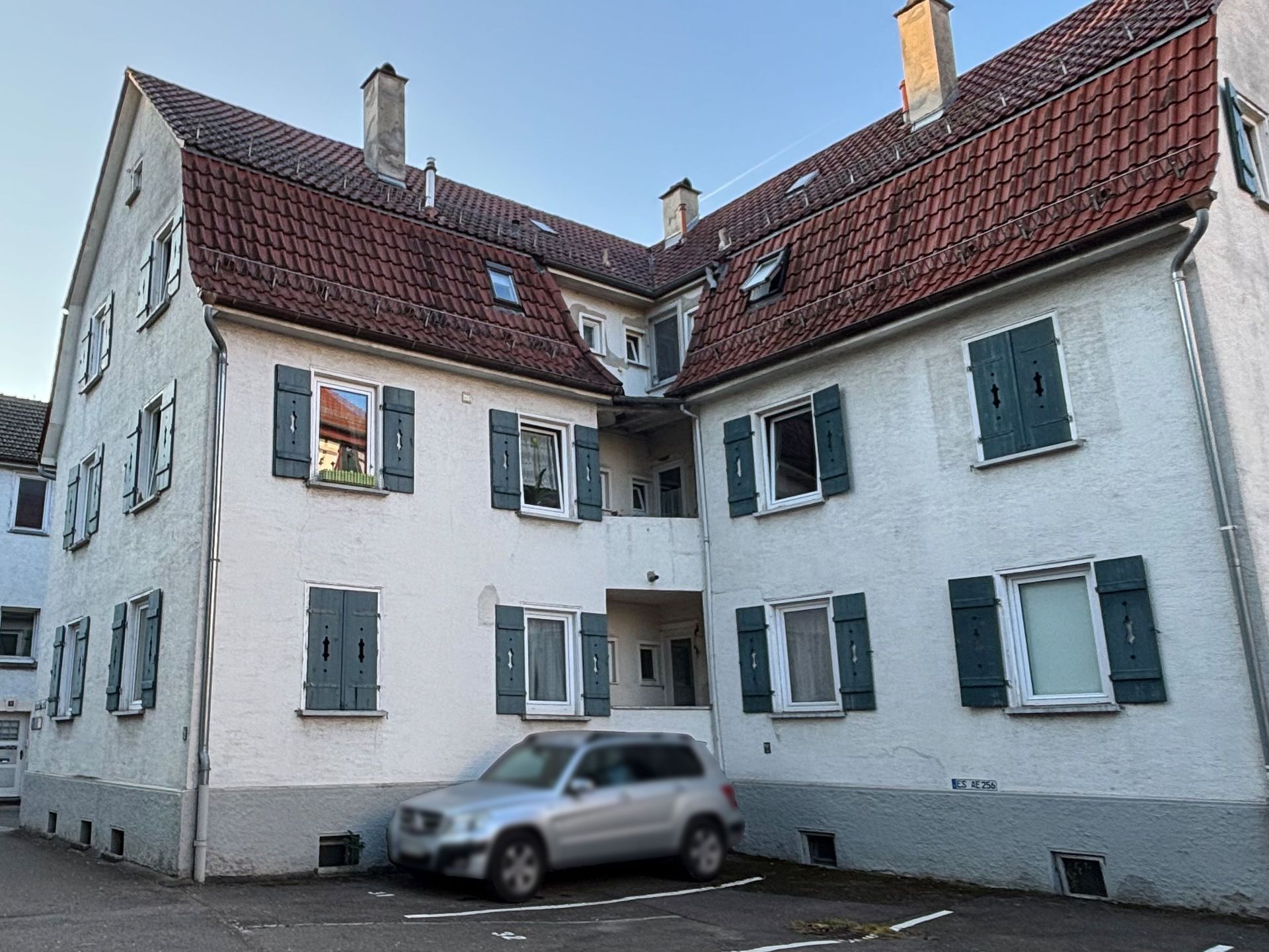 Das Bild zeigt ein weißes Mehrfamilienhaus mit einem traditionellen, roten Ziegeldach und grünen Fensterläden. Vor dem Haus befindet sich ein gepflasterter Hof mit einem geparkten Auto.