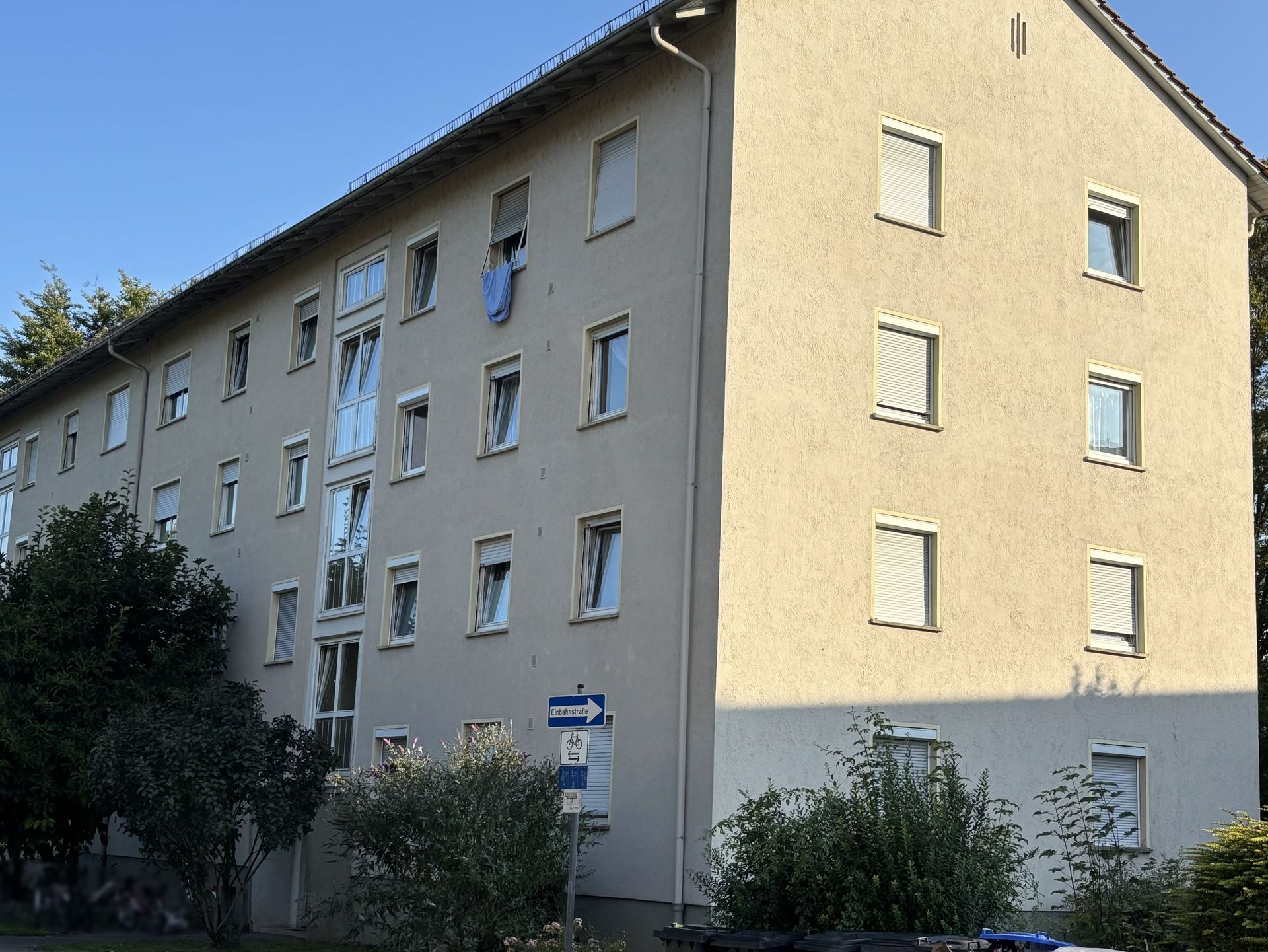 Das Bild zeigt ein Mehrfamilienhaus mit heller Fassade. Vor dem Haus stehen einige Bäume und Sträucher.