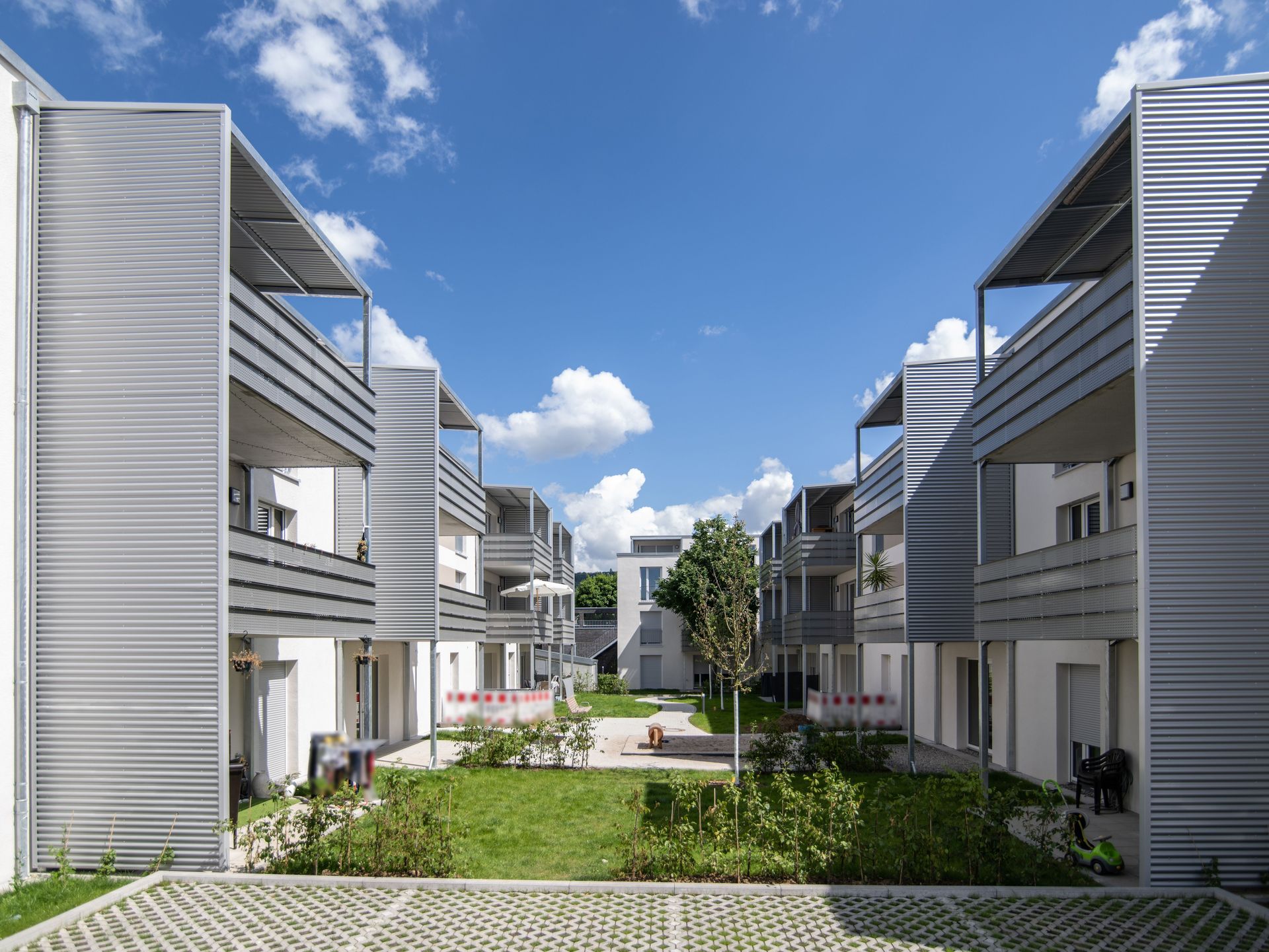 Das Bild zeigt den Blick durch zwei Mehrfamilienhäuser. Die Häuser haben eine helle Fassade und graue Balkone. Es sind Rasenflächen zu sehen mit Sträuchern und kleinen Bäumen.