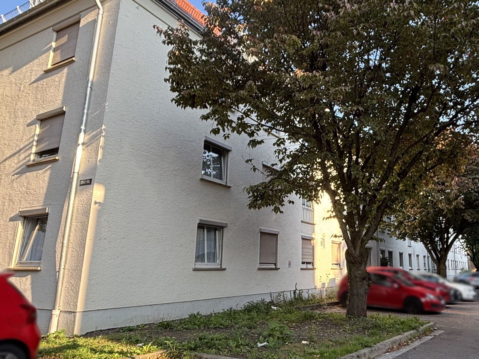 Das Bild zeigt ein Mehrfamilienhaus mit heller Fassade und mehreren Stockwerken. Vor dem Haus stehen Bäume und am Straßenrand parken Autos.