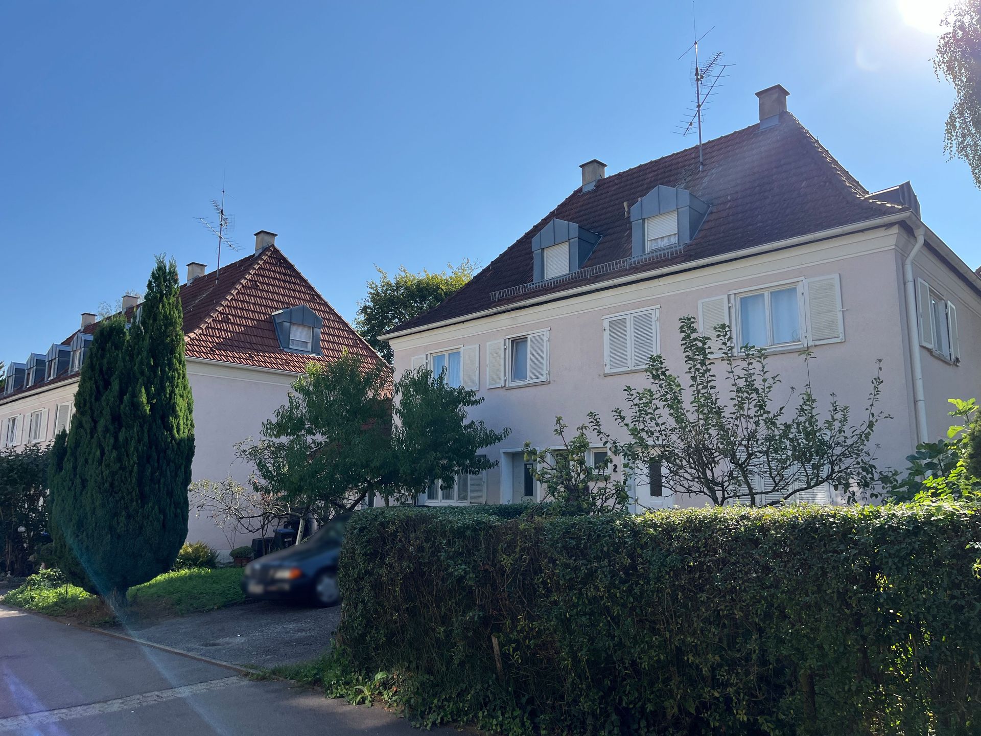 Das Bild zeigt zwei Häuser mit heller Fassade, Ziegeldächern und weißen Fensterläden. Vor den Häusern steht eine hohe Hecke und dahinter sind Bäume zu erkennen.