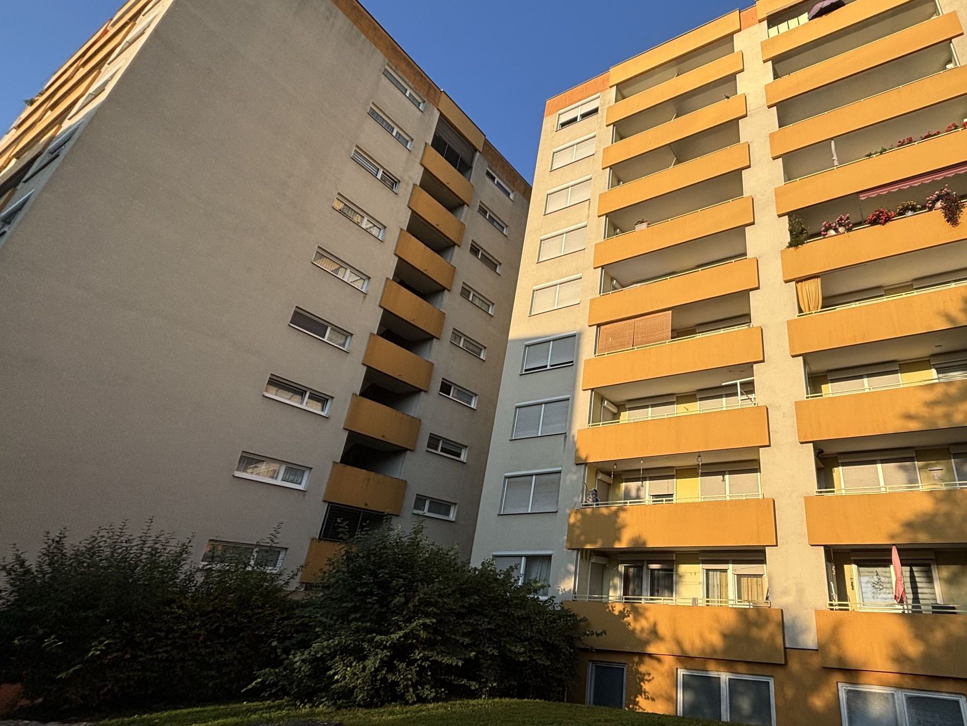 Das Bild zeigt zwei nebeneinander stehende Mehrfamilienhäuser mit auffälligen, orangefarbenen Balkonen. Zwischen den Gebäuden befindet sich etwas Grün und die Sonne scheint auf die Fassaden.