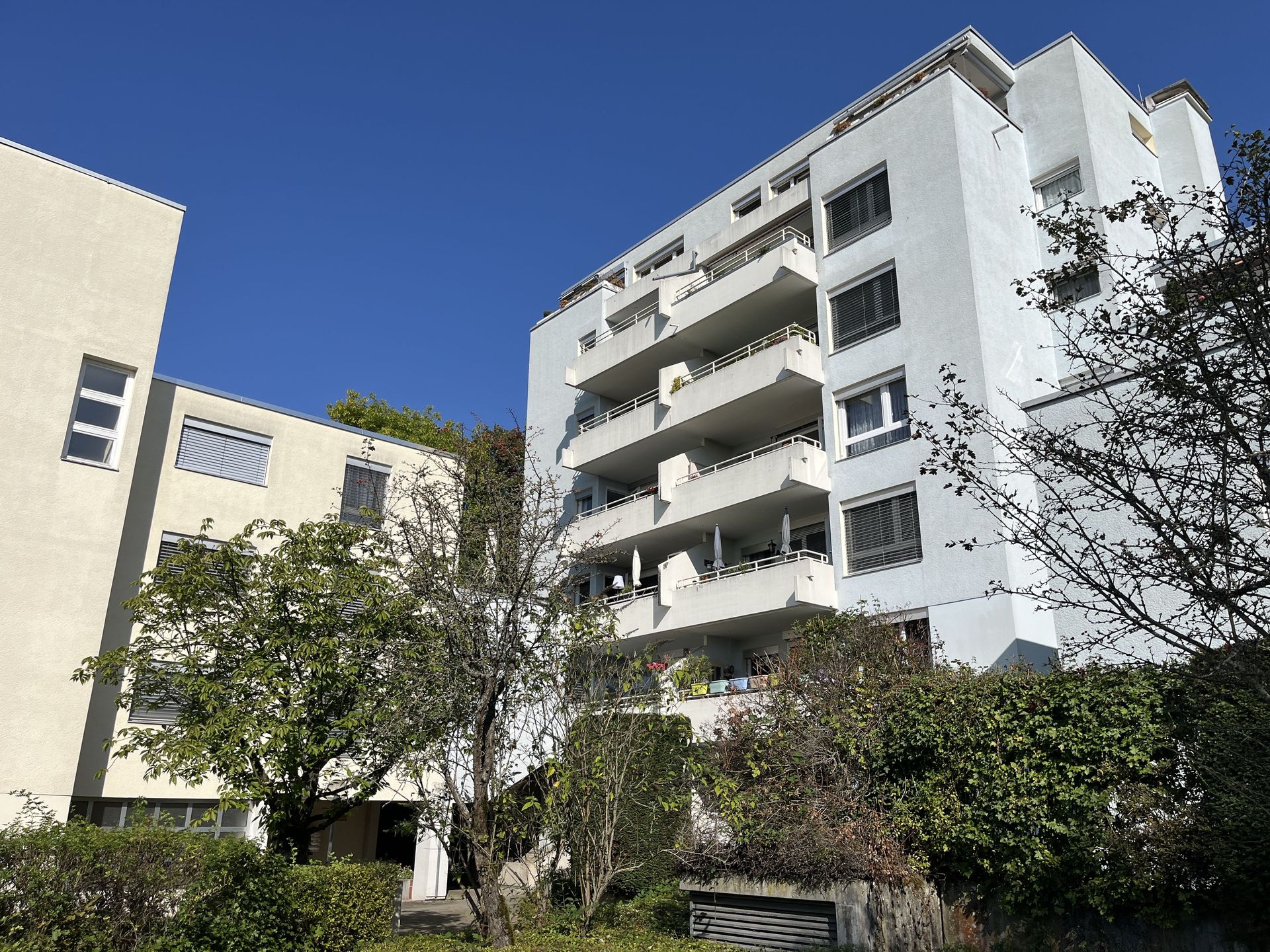 Das Bild zeigt ein Mehrfamilienhaus mit heller Fassade. Vor dem Haus befindet sich eine Grünfläche mit Büschen und Bäumen.