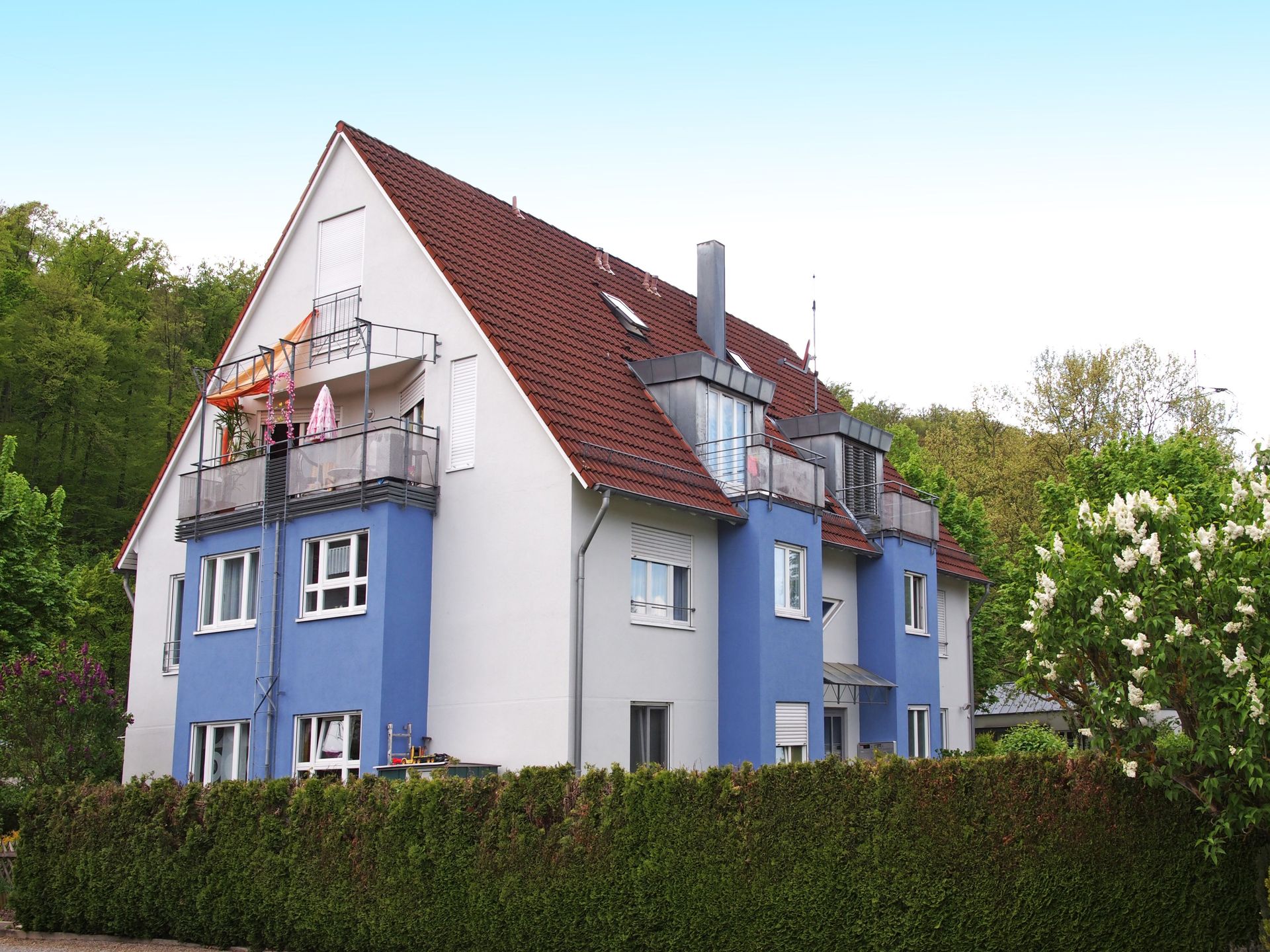 Das Bild zeigt ein Mehrfamilienhaus mit blau-weißer Fassade und einem roten Ziegeldach. Vor dem Haus befindet sich eine grüne Hecke.