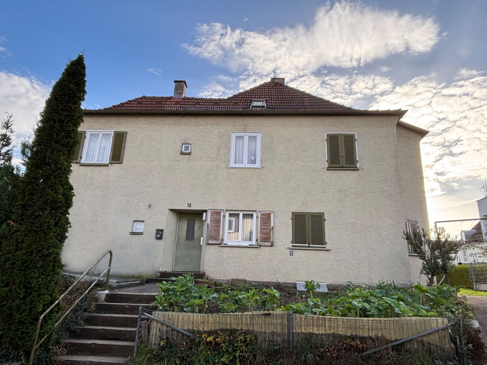 Das Bild zeigt ein Haus mit heller Fassade und braunen Fensterläden. Vor dem Haus ist ein Beet angelegt.