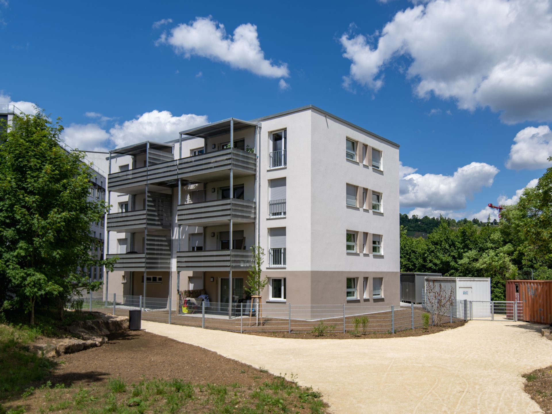 Das Bild zeigt ein modernes Mehrfamilienhaus mit weißer Fassade und grauen Balkonen. Um das Haus herum befindet sich eine Grünfläche mit Bäumen und ein Weg führt zum Gebäude.