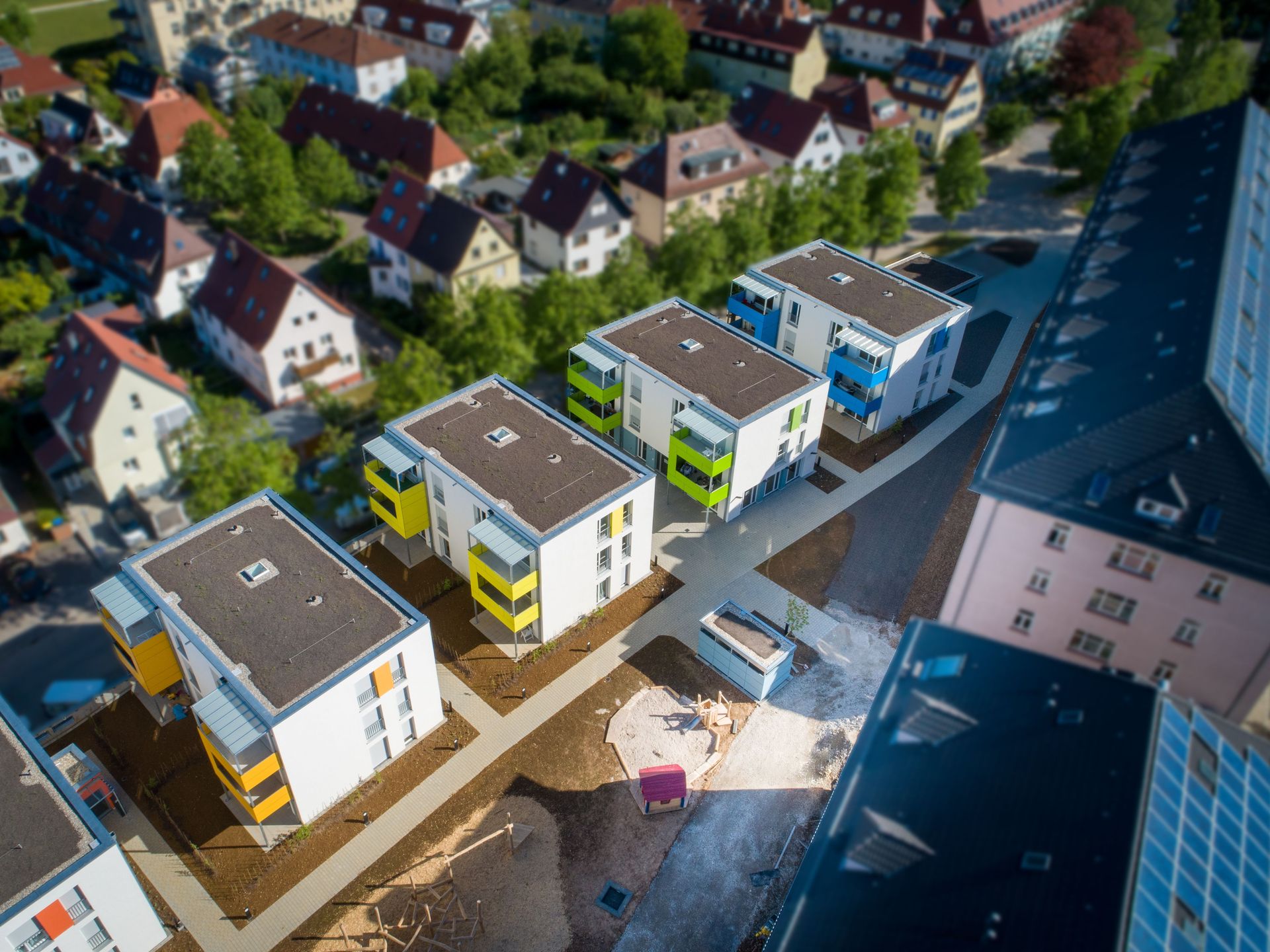 Das Bild zeigt eine moderne Wohnsiedlung mit mehreren Mehrfamilienhäusern, deren Balkone in verschiedenen Farben – gelb, grün und blau – gestaltet sind. Die Häuser sind von Straßen und Wegen umgeben und fügen sich in ein bestehendes Wohngebiet mit verschiedenen Gebäuden und viel Grün ein.