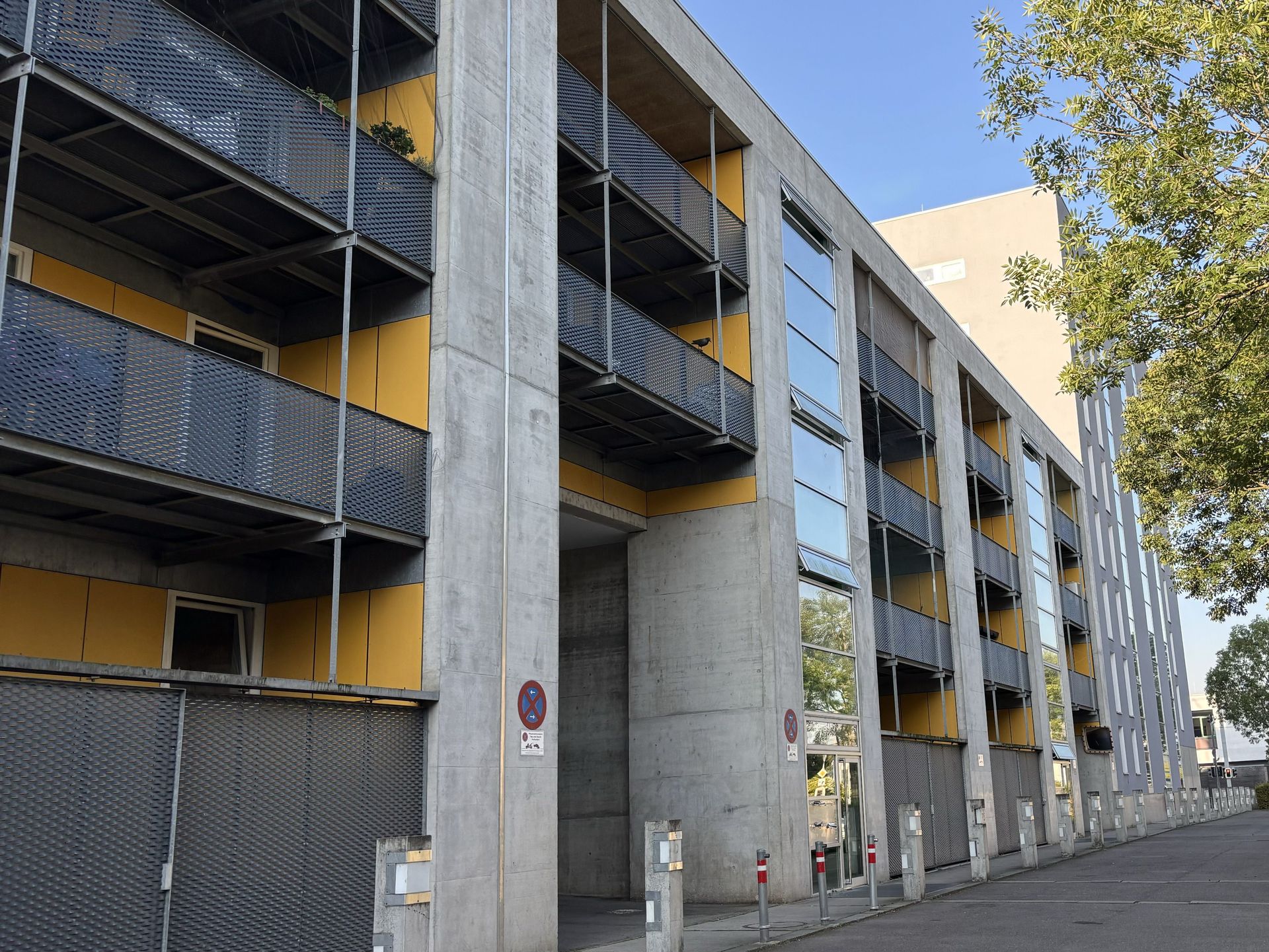 Das Bild zeigt ein modernes Gebäude mit grauer Betonfassade und gelben Elementen. Das Gebäude hat mehrere Stockwerke mit Balkonen und großen Fenstern.