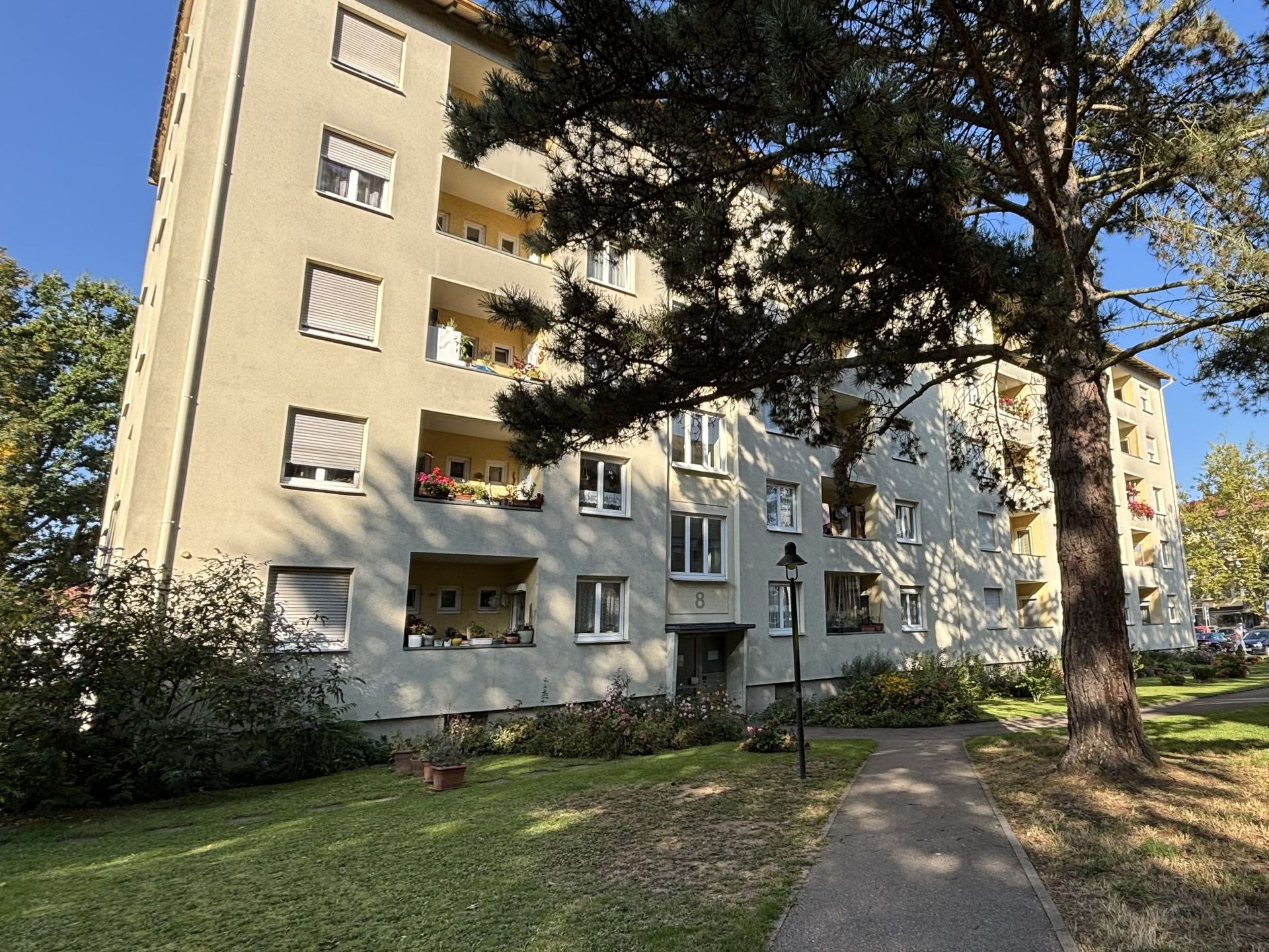 Das Bild zeigt ein großes Mehrfamilienhaus mit heller Fassade.  Vor dem Haus befindet sich eine Grünfläche mit Bäumen und ein Weg führt zum Eingang.