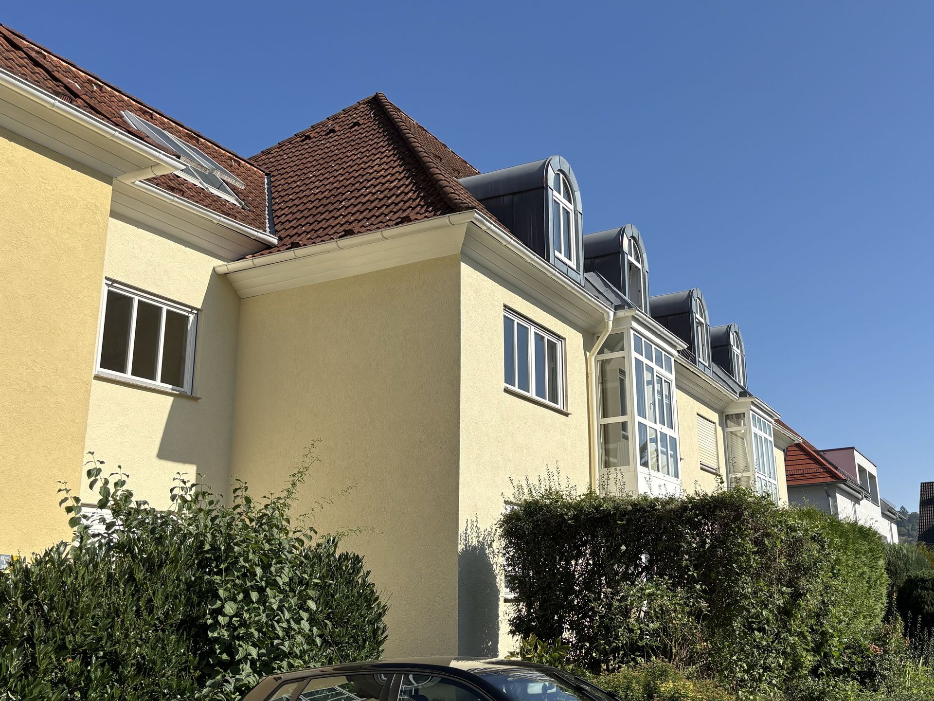 Das Bild zeigt ein Haus mit hellgelber Fassade und markanten, abgerundeten Dachgauben. Vor den Häusern befindet sich eine Hecke und im Hintergrund sind weitere Gebäude zu sehen.