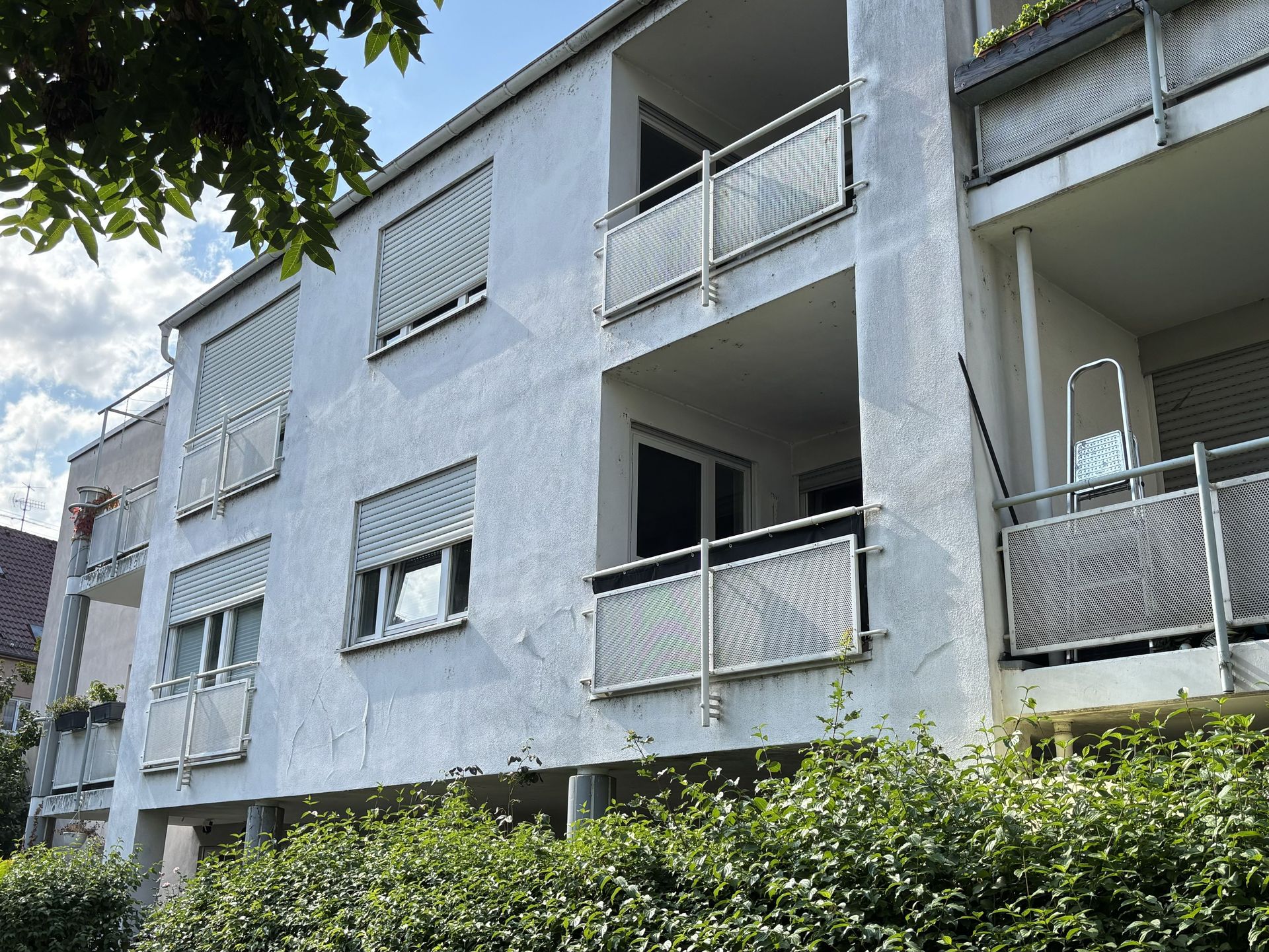 Das Bild zeigt ein Mehrfamilienhaus mit weißer Fassade, das von Büschen und Bäumen umgeben ist. Die Balkone des Hauses haben unterschiedliche Größen und Geländer.