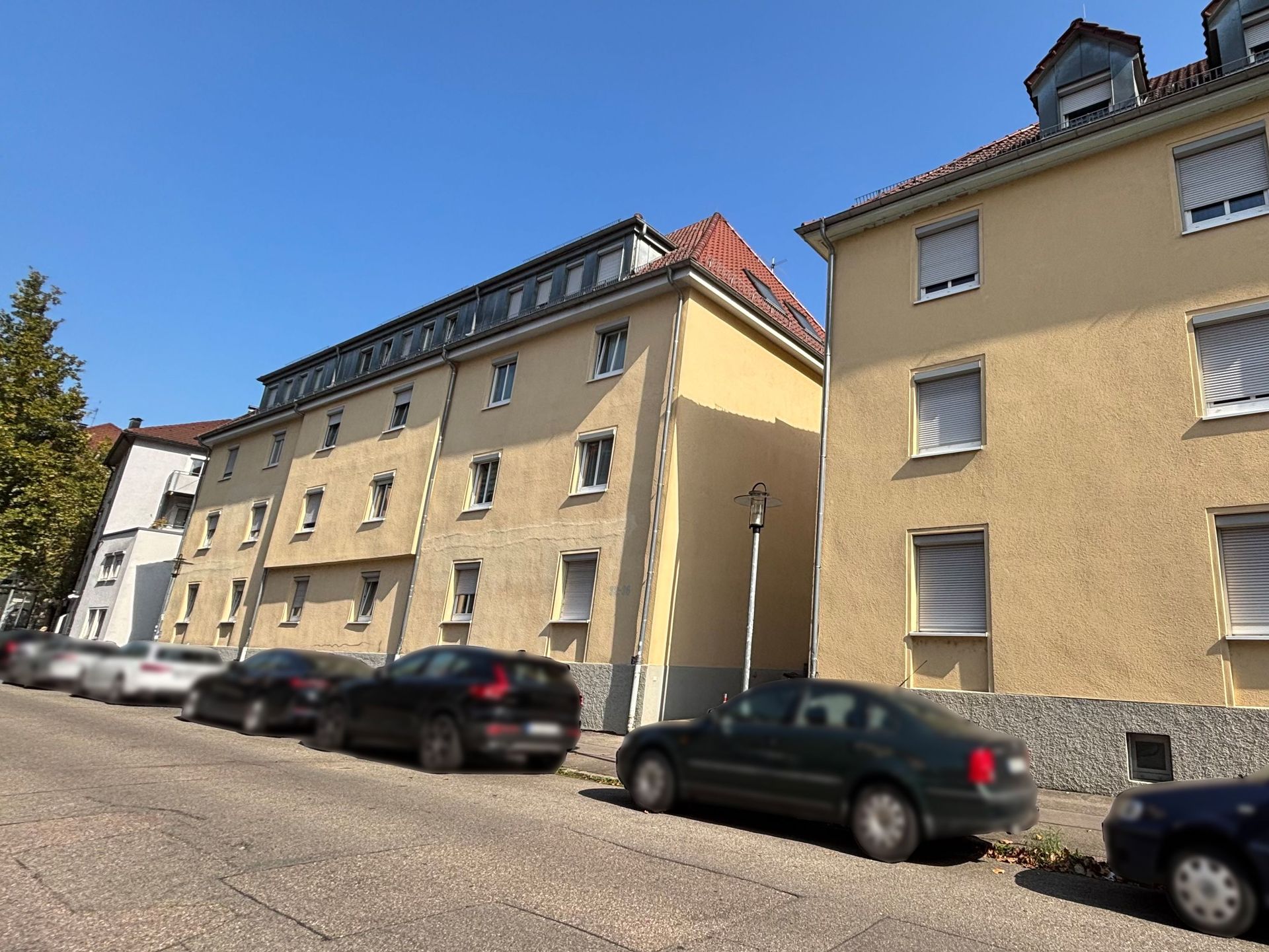 Das Bild zeigt zwei nebeneinander stehende Mehrfamilienhäuser mit hellgelber Fassade. Vor den Häusern parken mehrere Autos am Straßenrand und im Hintergrund sind Bäume zu sehen.