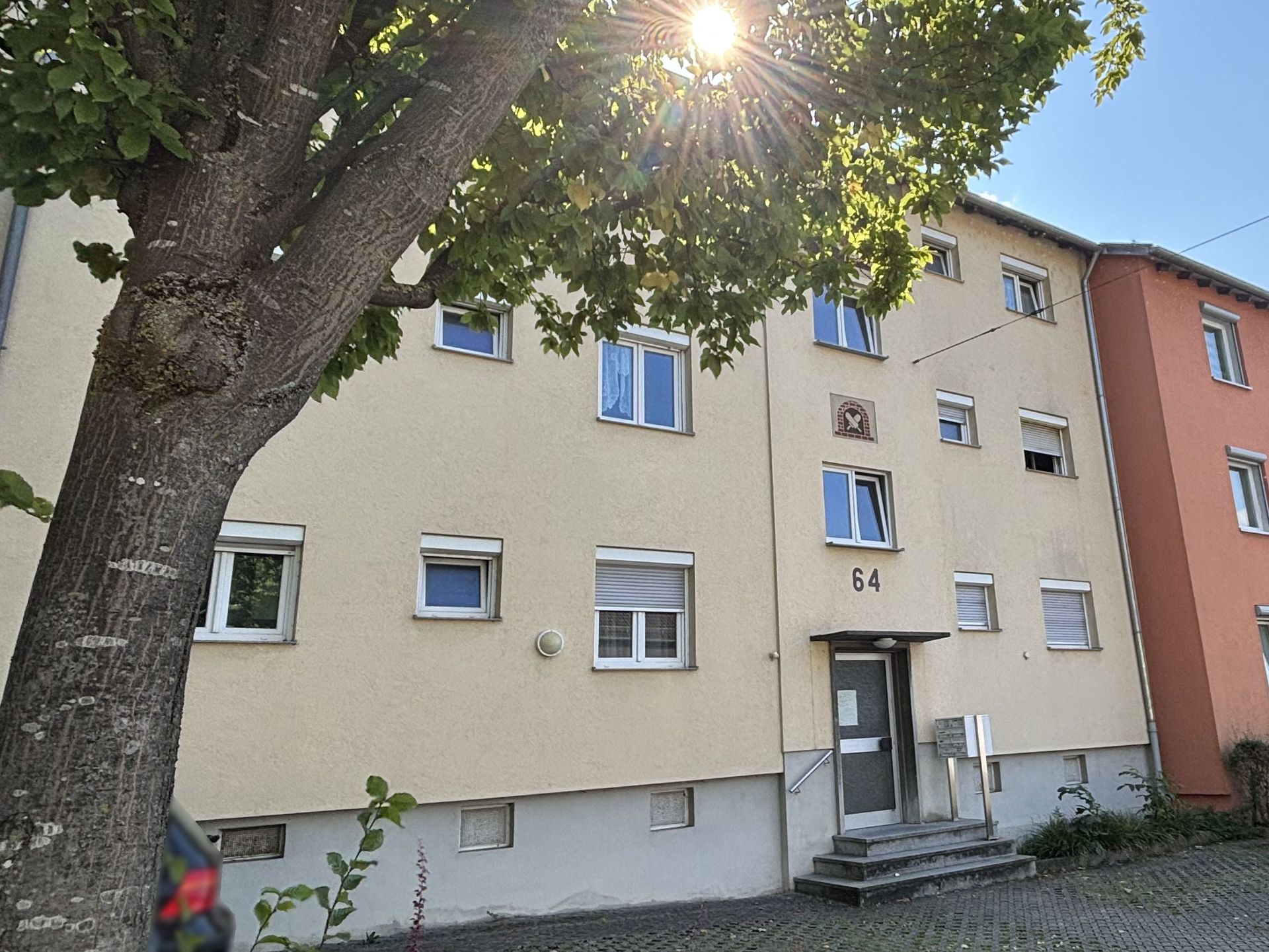 Das Bild zeigt ein Mehrfamilienhaus mit hellgelber Fassade und unterschiedlich angeordneten Fenstern. Zwischen den Fenstern über der Eingangstür schmücken Abbildungen den Haus. Links im Bild steht ein großer Baum, durch den die Sonne hindurch scheint.
