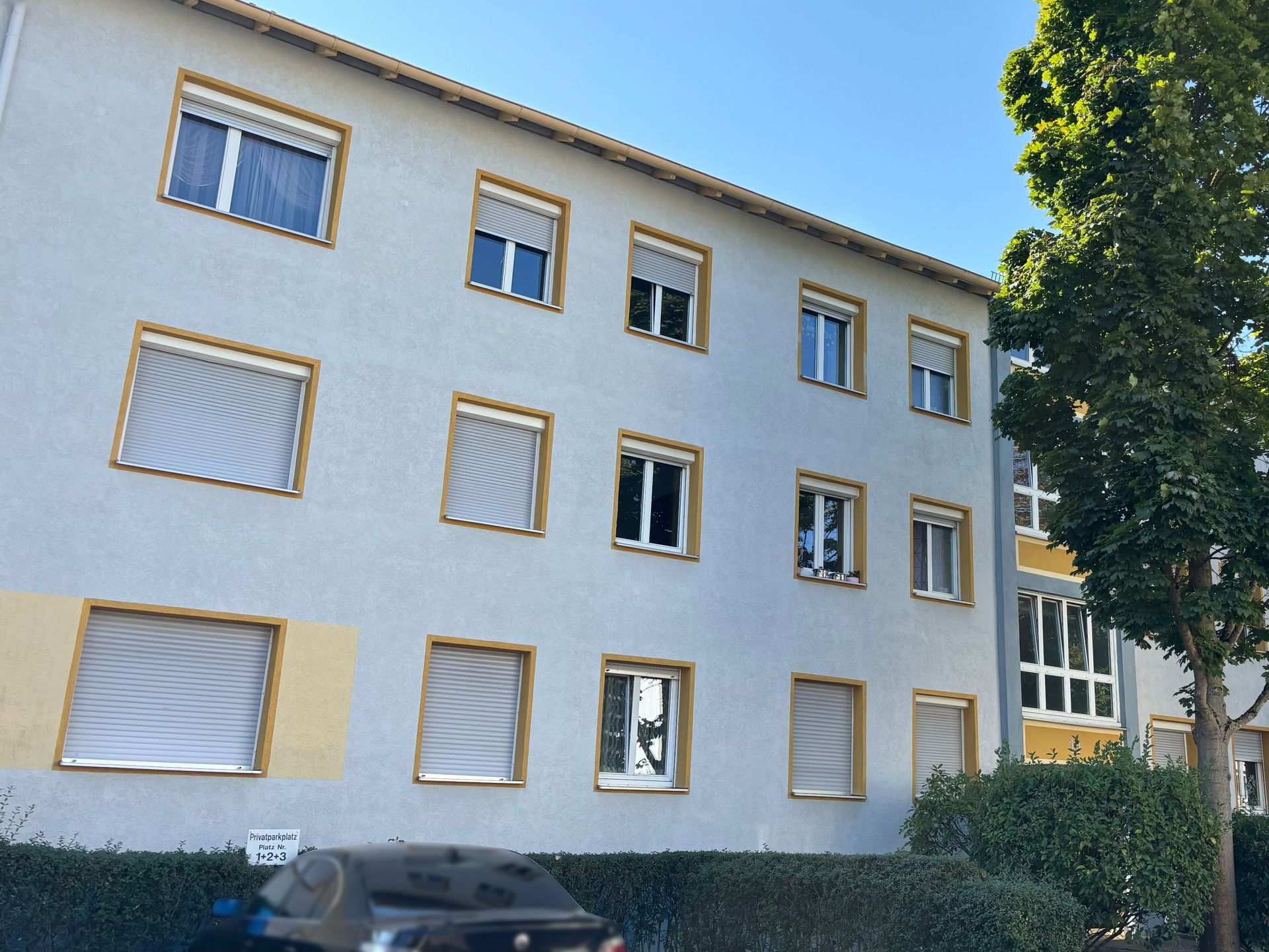 Das Bild zeigt ein Mehrfamilienhaus mit heller Fassade und gelben Fensterrahmen. Vor dem Haus parkt ein Auto und rechts im Bild steht ein Baum.