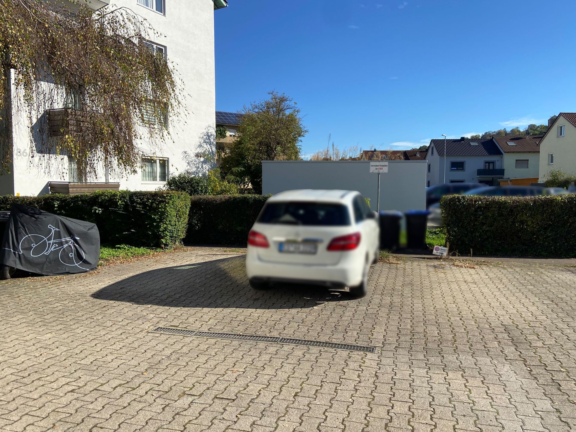 Das Bild zeigt einen Parkplatz mit einem geparkten Auto. Der Parkplatz ist von einer Hecke umgeben und im Hintergrund sind weitere Gebäude zu sehen.