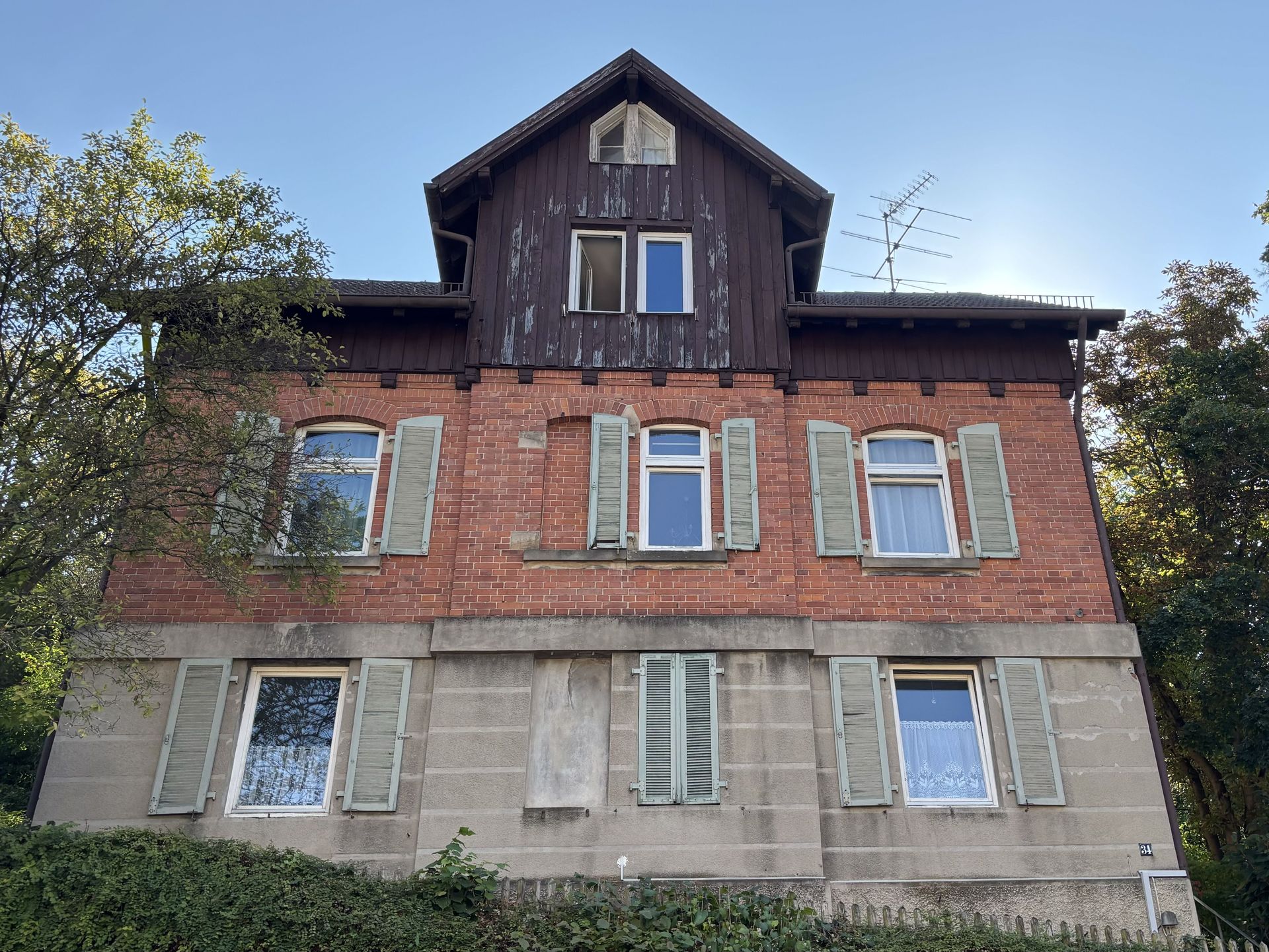 Das Bild zeigt ein mehristöckiges Haus mit roter Klinkerfassade im oberen Bereich und verputztem Sockel im unteren Bereich. Das Haus hat ein dunkles Dach und helle Fensterläden.