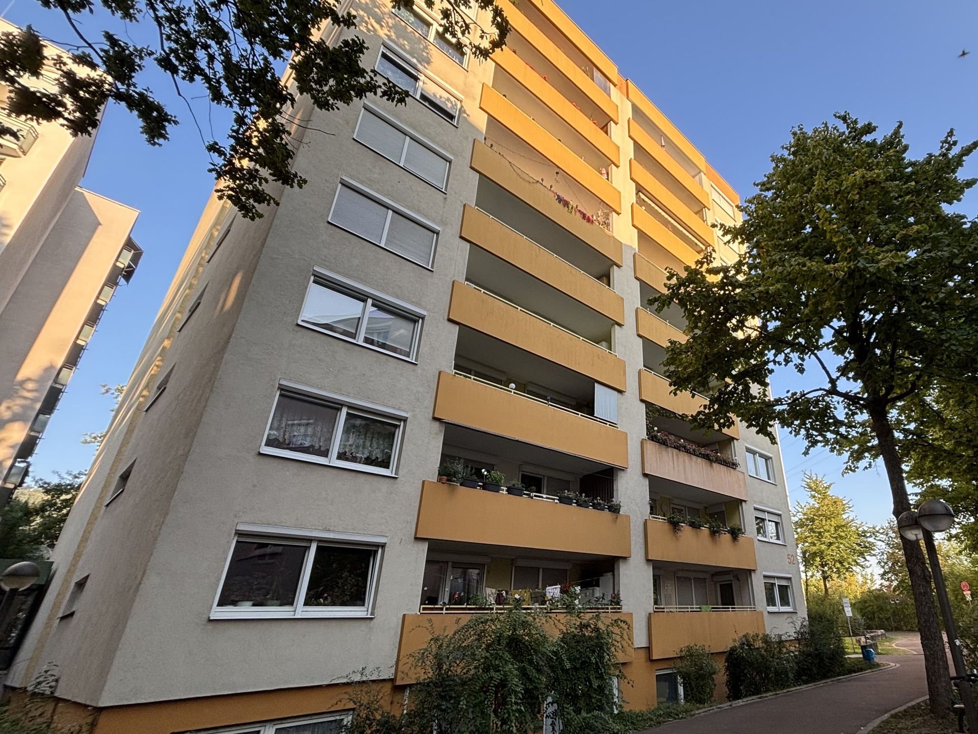 Das Bild zeigt ein Mehrfamilienhaus mit heller Fassade und auffälligen, orangefarbenen Balkonen, die sich über die gesamte Höhe des Gebäudes erstrecken.  Umgeben ist das Haus von Bäumen und Grünflächen, und ein Weg führt am Gebäude vorbei.