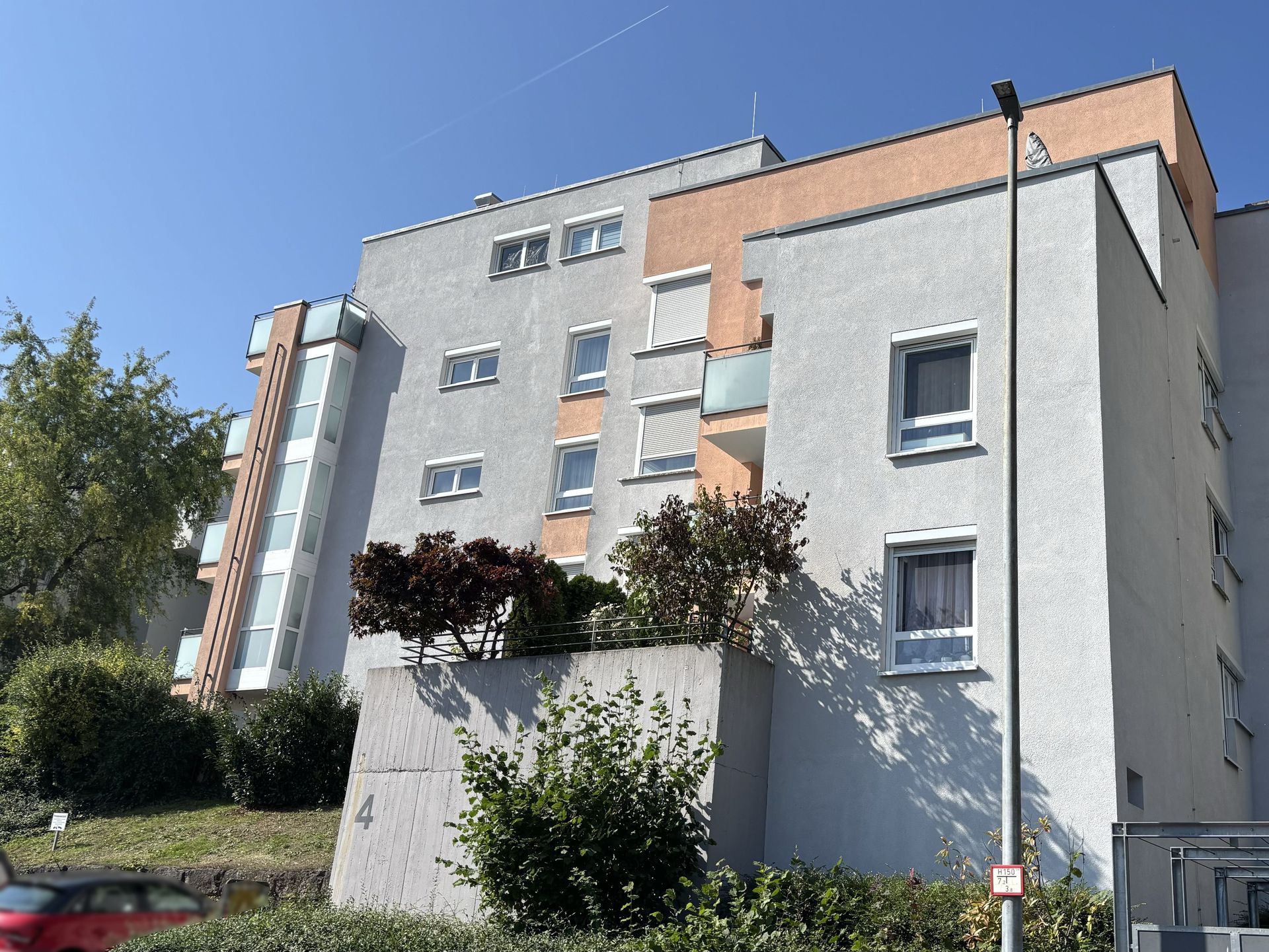Das Bild zeigt ein Mehrfamilienhaus mit heller Fassade und einem orangefarbenen Bereich im oberen Teil. Vor dem Haus stehen einige Bäume und Sträucher.