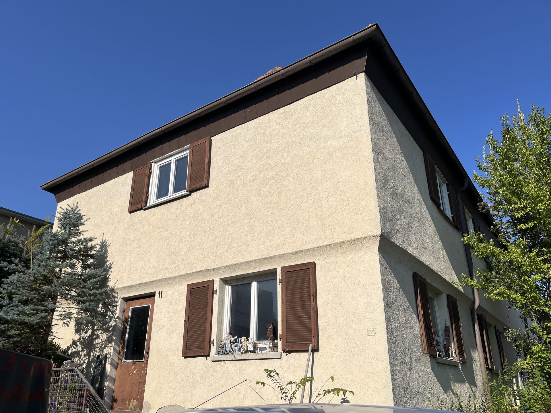 Das Bild zeigt ein helles Haus mit braunem Dach und braunen Fensterläden.  Eine Außentreppe führt zum Hauseingang und vor dem Haus steht ein Baum.