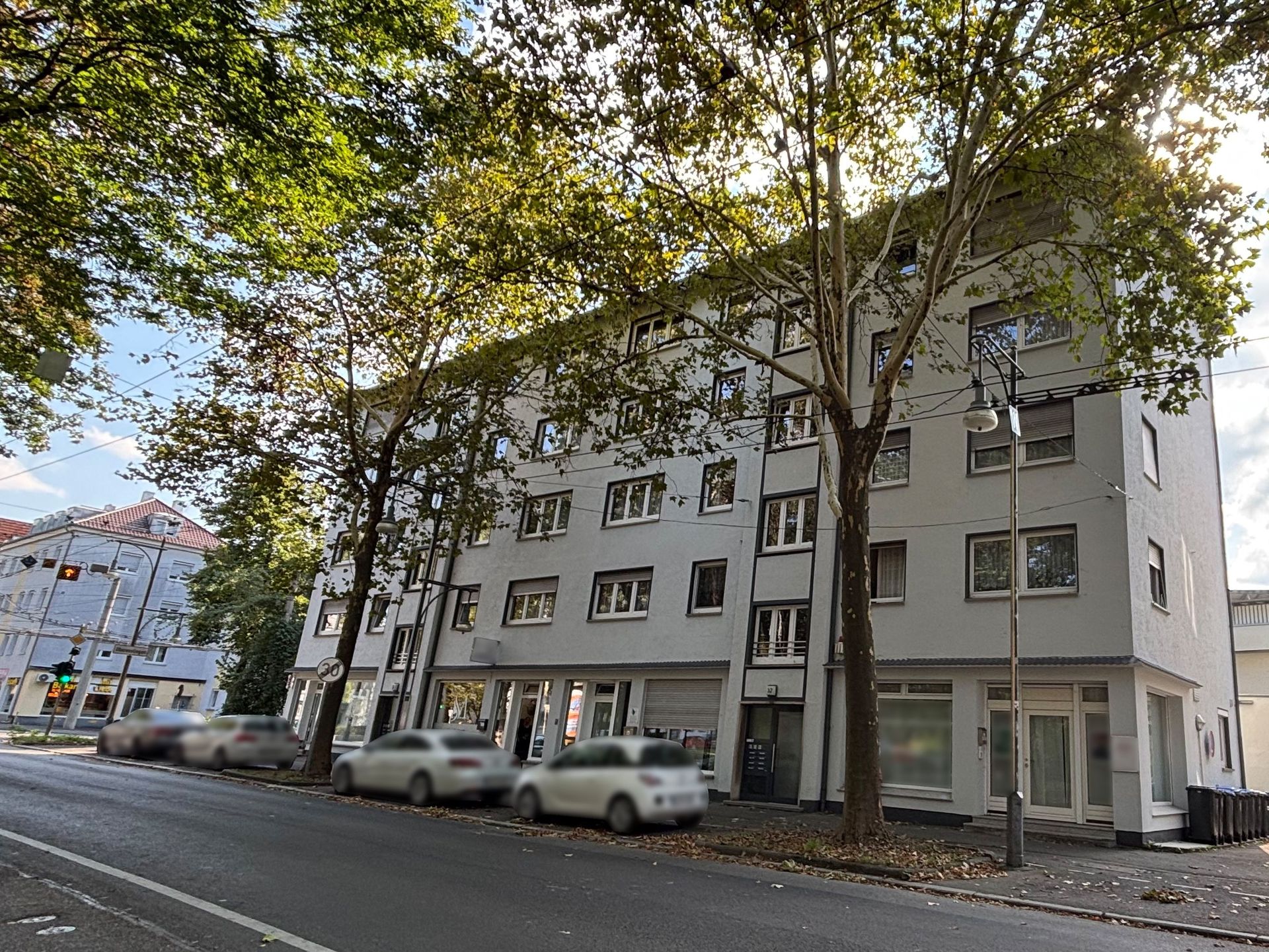 Das Bild zeigt ein Mehrfamilienhaus mit heller Fassade und mehreren Stockwerken, vor dem Autos am Straßenrand geparkt sind. Hohe Bäume säumen den Gehweg und spenden Schatten auf das Gebäude und die Straße.
