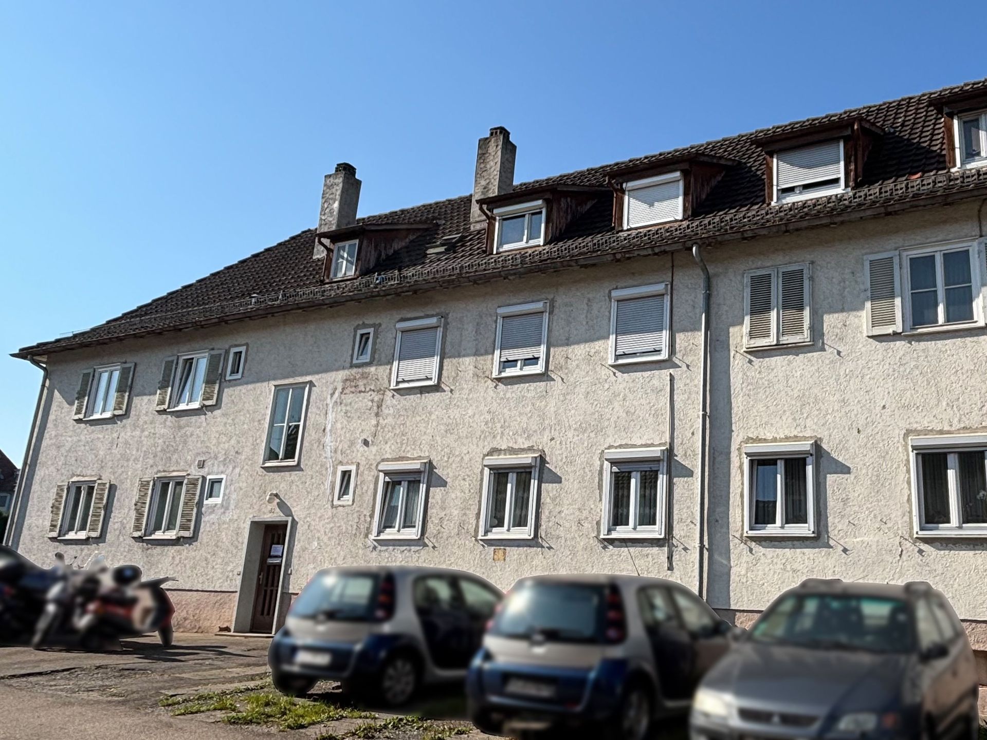 Das Bild zeigt ein Mehrfamilienhaus mit heller Fassade, dunklem Dach und einer Reihe von Dachgauben. Vor dem Haus befinden sich ein Parkplatz mit mehreren Autos und ein Motorrad.