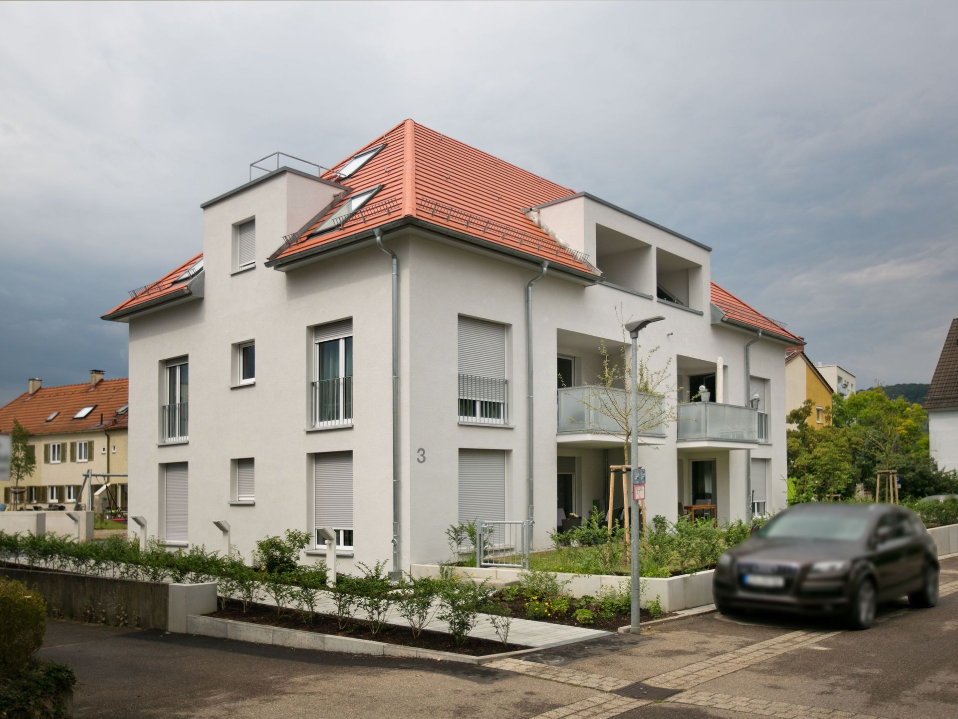 Das Bild zeigt ein modernes Mehrfamilienhaus mit weißer Fassade und rotem Ziegeldach.  Vor dem Haus befindet sich ein kleiner Garten mit Sträuchern und ein gepflasterter Weg führt zum Eingang. Daneben steht ein Auto.