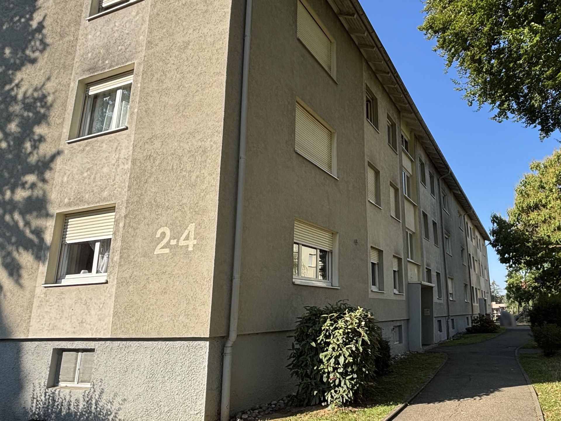 Das Bild zeigt ein Mehrfamilienhaus mit beiger Fassade. Entlang des Gebäudes führt ein schmaler Weg lang , der von Bäumen und Sträuchern gesäumt wird.