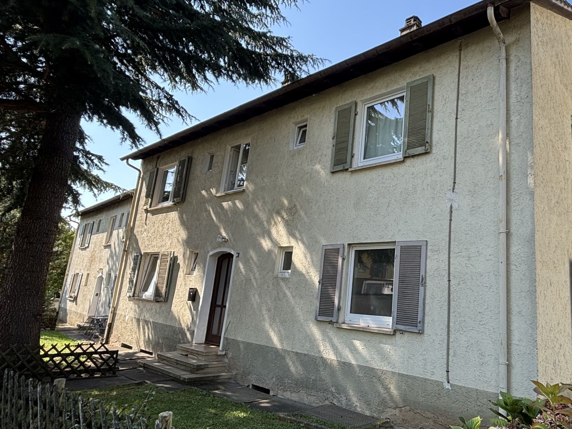 Das Bild zeigt zwei Häuser mit heller Fassade und Fensterläden. Links im Bild ist ein großer Baum zu sehen, der Schatten auf das Haus wirft.