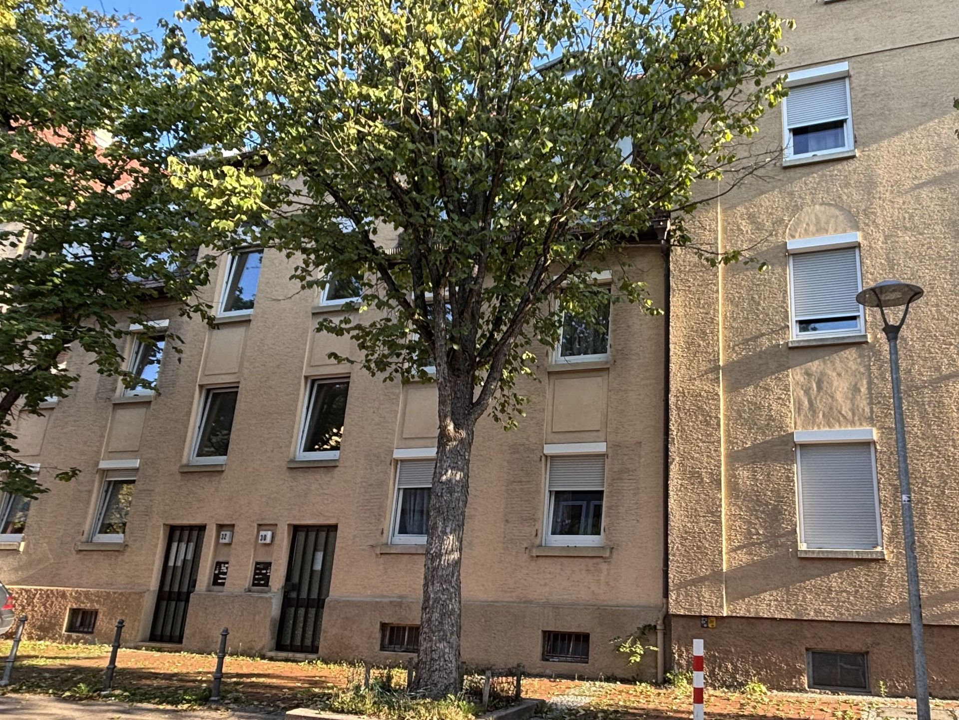Das Bild zeigt ein Mehrfamilienhaus mit heller Fassade und mehreren Stockwerken. Vor dem Haus steht ein großer Baum, der Schatten auf das Gebäude wirft.