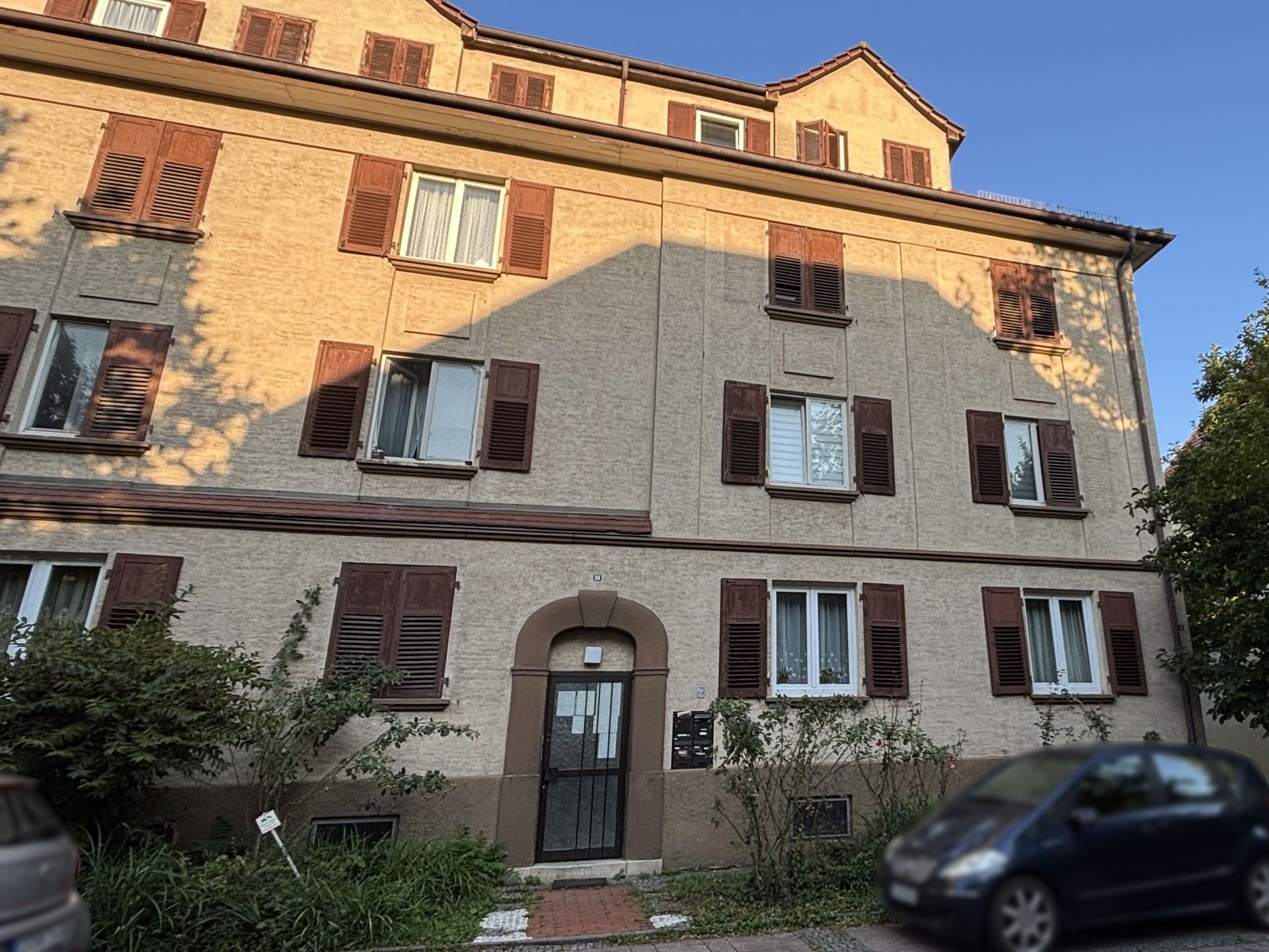Das Bild zeigt ein Mehrfamilienhaus mit heller Fassade und braunen Fensterläden. Vor dem Haus parkt ein Auto am Straßenrand und im Vordergrund sind Büsche zu sehen.