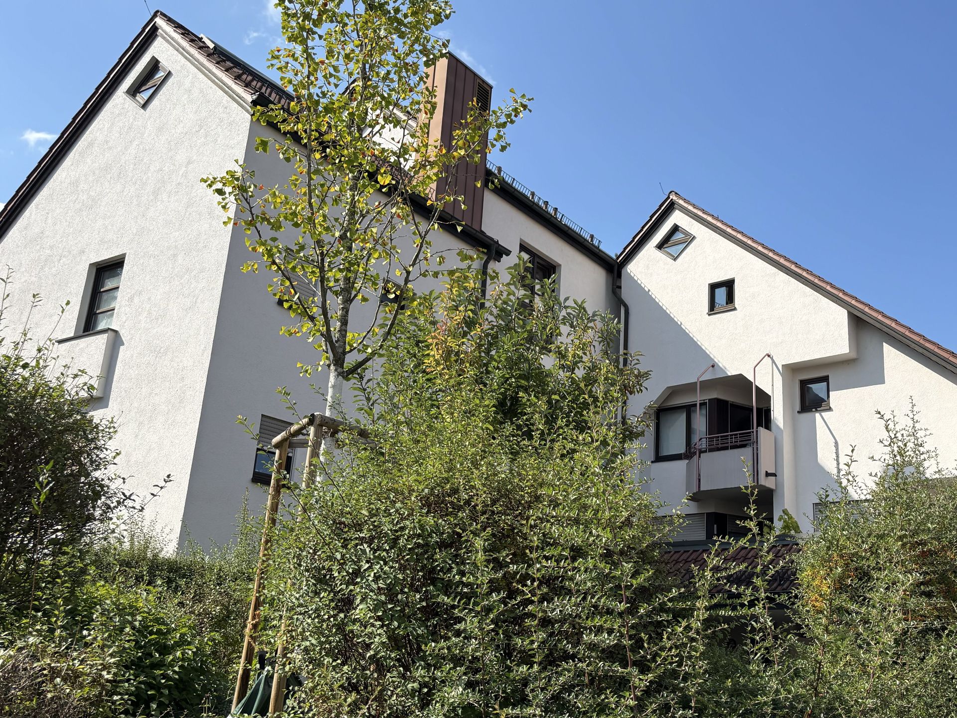 Das Bild zeigt ein modernes Mehrfamilienhaus mit weißer Fassade und dunklem Dach. Vor dem Haus befindet sich Bäume und Sträucher.