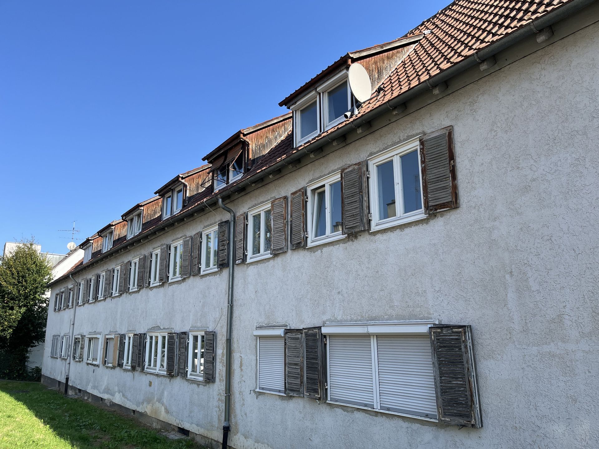 Das Bild zeigt ein langgezogenes Mehrfamilienhaus mit heller Fassade, braunem Ziegeldach und Fensterläden.