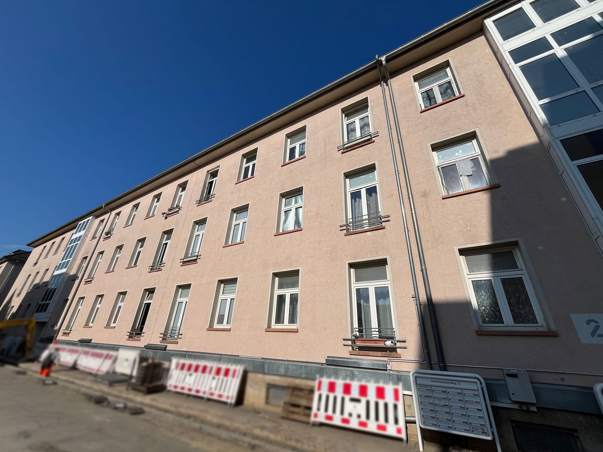 Das Bild zeigt ein Mehrfamilienhaus mit heller Fassade und vielen gleichförmigen Fenstern, an dem ein moderner, verglaster Anbau zu sehen ist. Vor dem Haus befinden sich Baustellenabsperrungen.