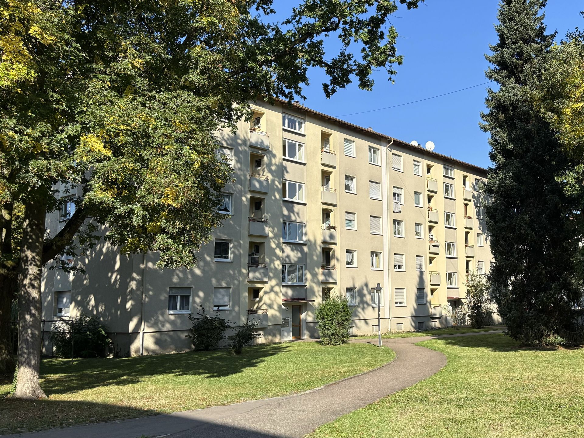 Das Bild zeigt ein großes Mehrfamilienhaus mit heller Fassade, umgeben von Bäumen. Ein Weg führt durch die Grünfläche vor dem Haus zum Eingang.