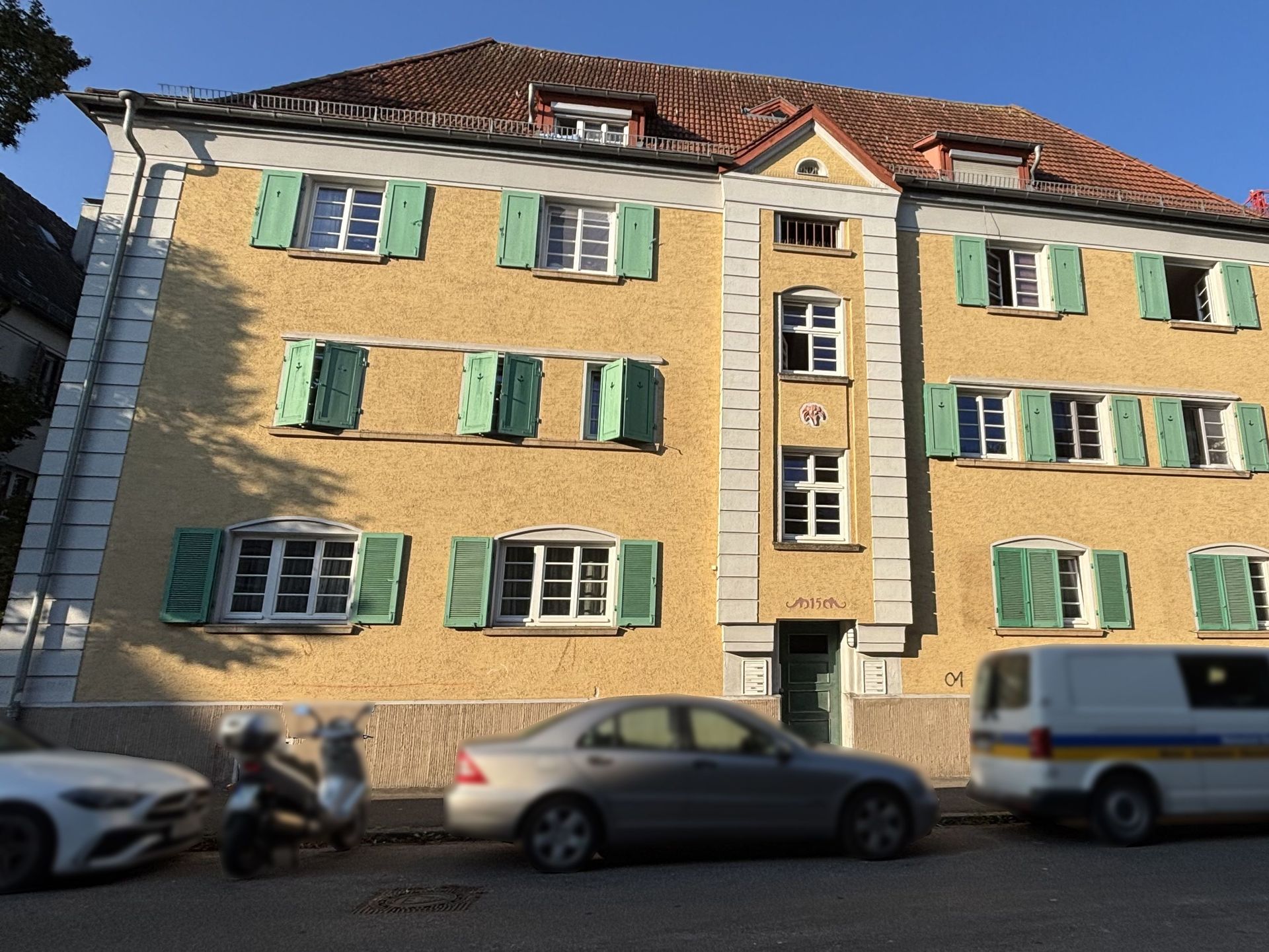 Das Bild zeigt ein Mehrfamilienhaus mit hellgelber Fassade und grünen Fensterläden. Vor dem Haus parken mehrere Fahrzeuge.