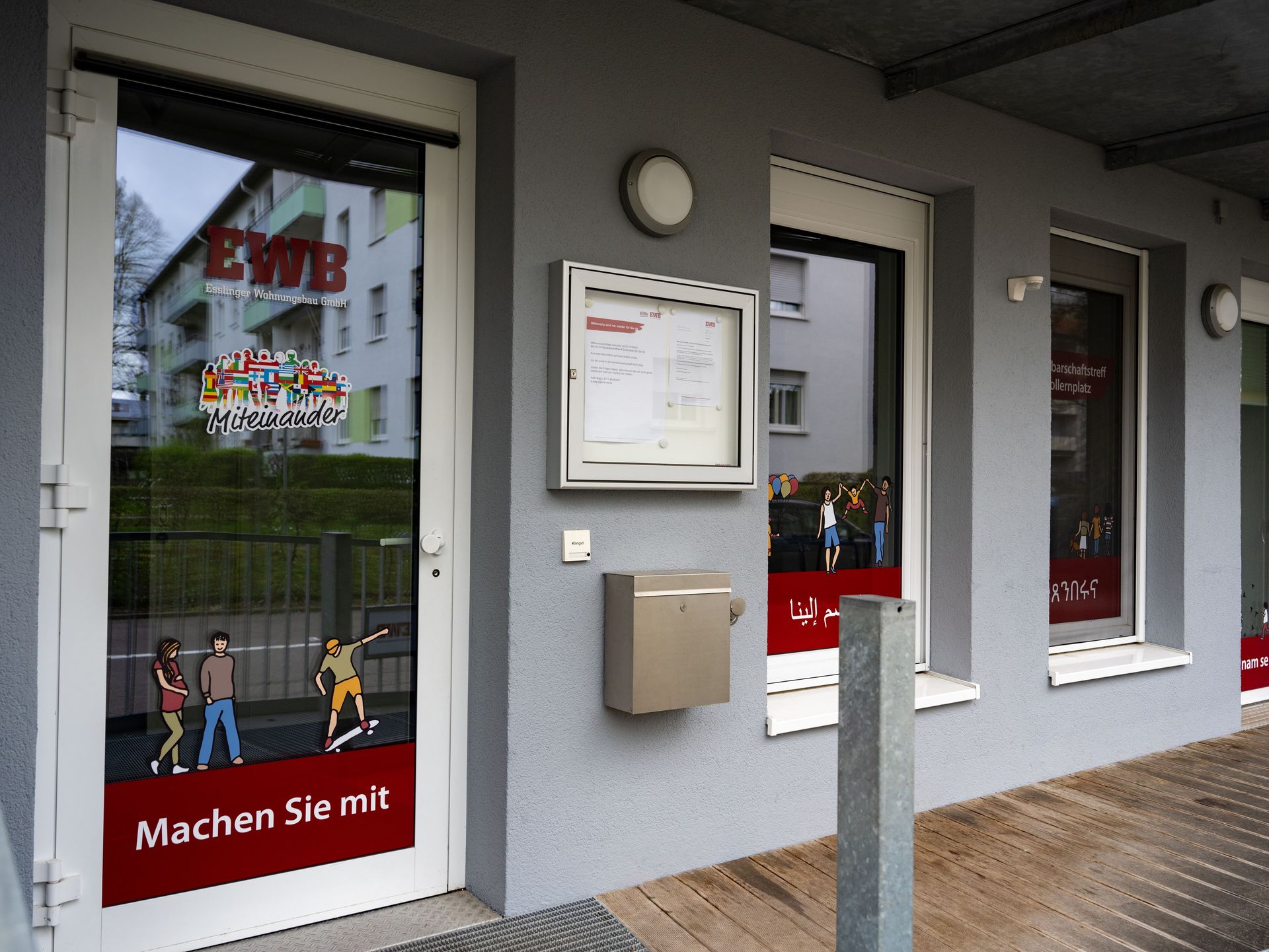 Das Bild zeigt einen Eingangsbereich. Die Fenster sind mit Gestaltungselementen beklebt und man sieht einen Briefkasten und einen Schaukasten, in dem Zettel hängen.