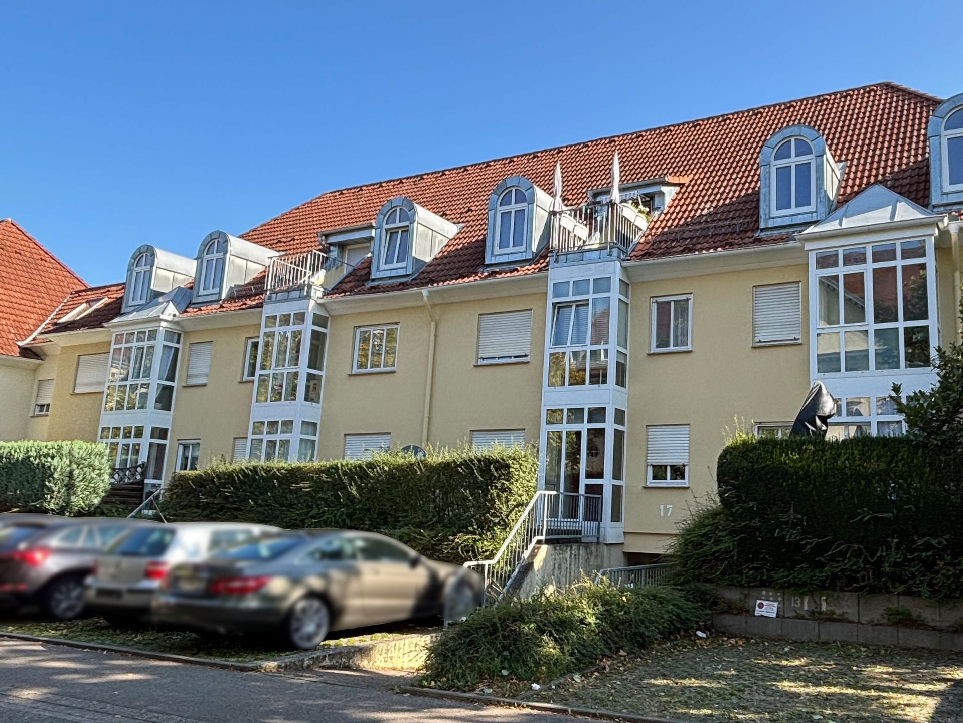 Das Bild zeigt ein Mehrfamilienhaus mit hellgelber Fassade, rotem Ziegeldach und auffälligen, abgerundeten Dachgauben. Vor dem Haus befinden sich Parkplätze, auf denen mehrere Autos abgestellt sind, und eine grüne Hecke.