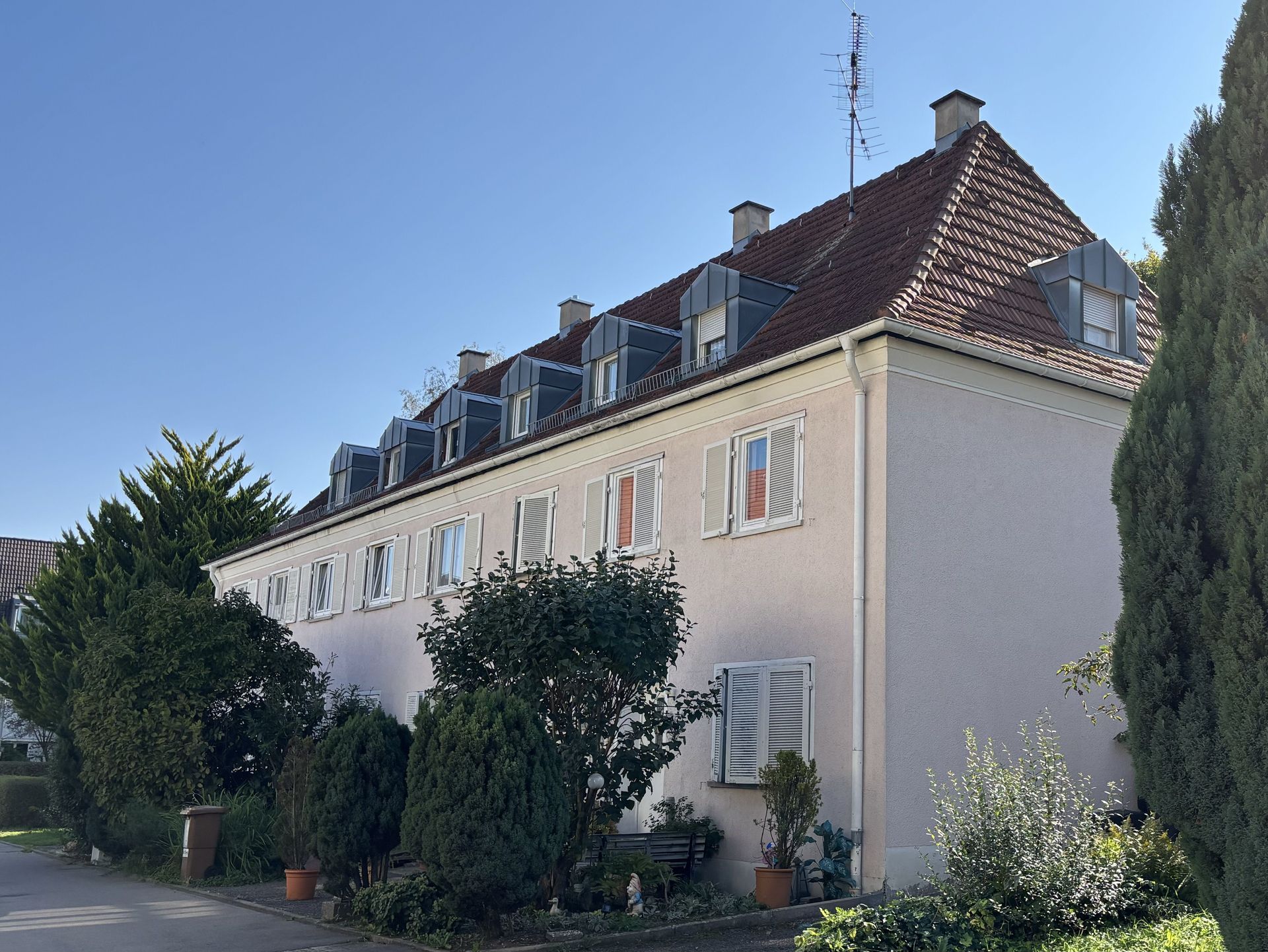 Das Bild zeigt ein Reihenhaus mit heller Fassade, Ziegeldach und Dachgauben.  Vor dem Haus befindet sich ein kleiner Vorgarten mit Sträuchern, Bäumen und Blumentöpfen.