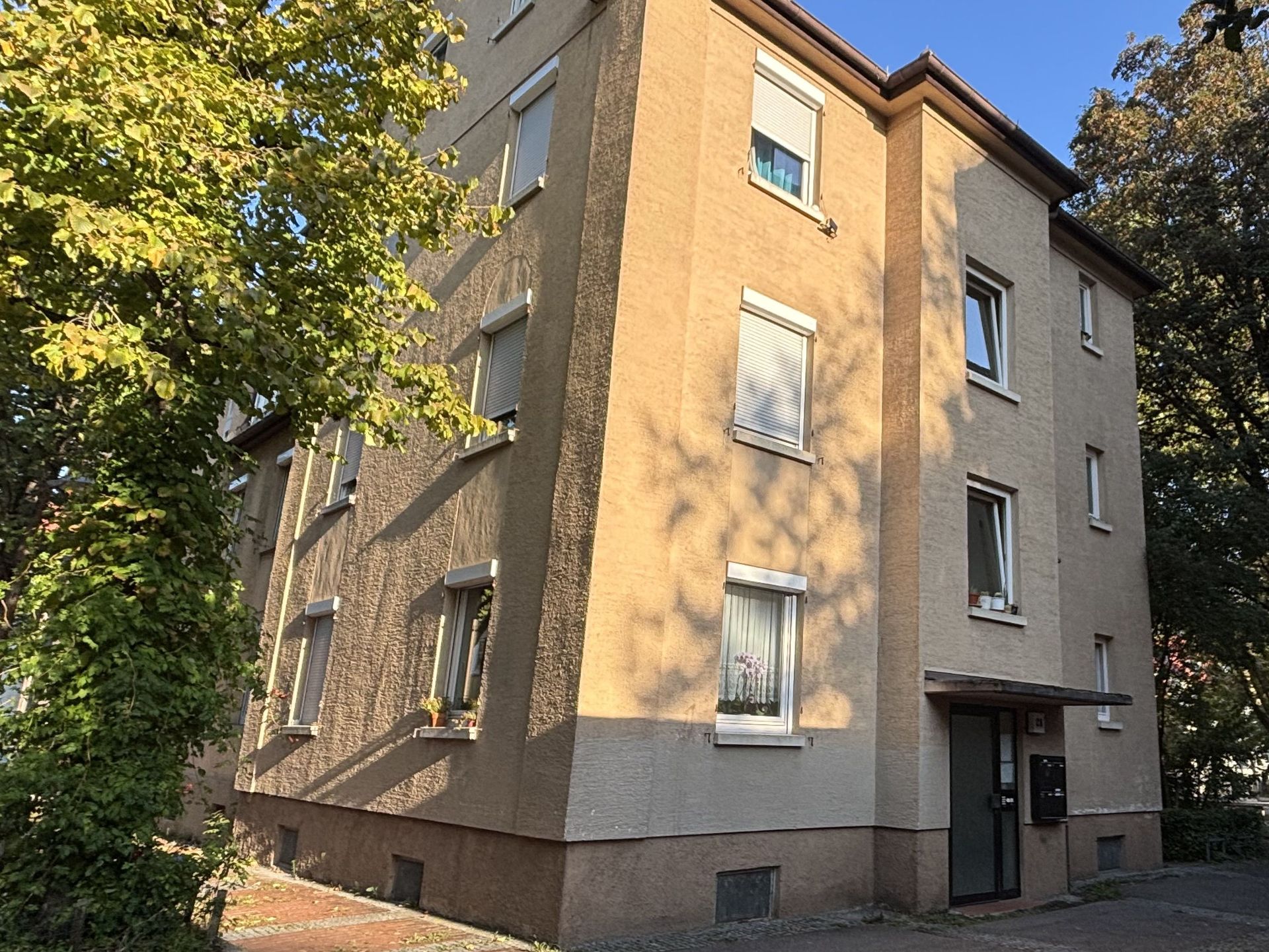 Das Bild zeigt ein Mehrfamilienhaus mit heller Fassade. Vor dem Haus stehen Bäume, die Schatten auf das Gebäude werfen.