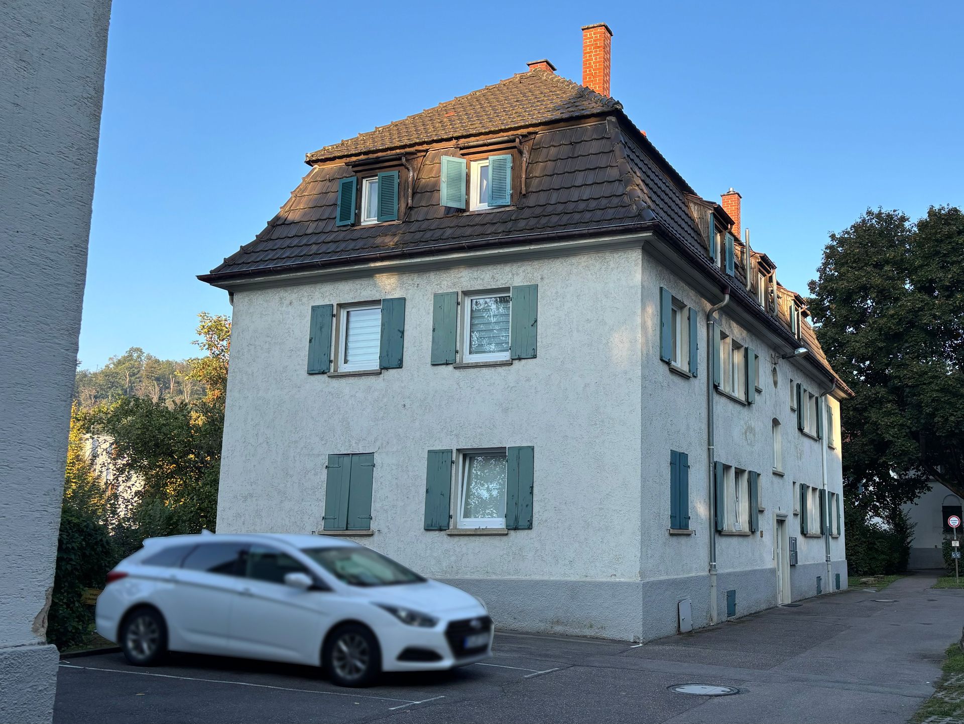 Das Bild zeigt ein mehrstöckiges Mehrfamilienhaus mit weißer Fassade und grünen Fensterläden.  Vor dem Haus befindet sich ein geparktes Auto und im Hintergrund sind Bäume zu sehen.