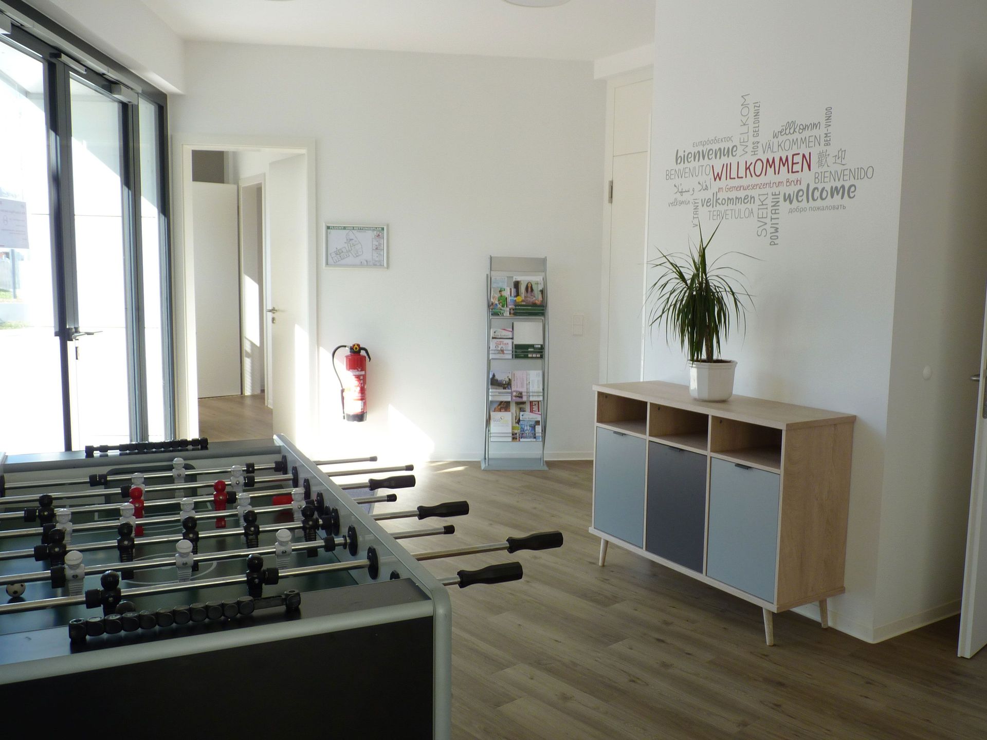 Das Bild zeigt eine Innenaufnahme des Gebäudes. Man sieht einen Tischkicker, eine Kommode mit Pflanze und ein Regal mit Broschüren. Auf der Wand steht Willkommen auf mehreren Sprachen.
